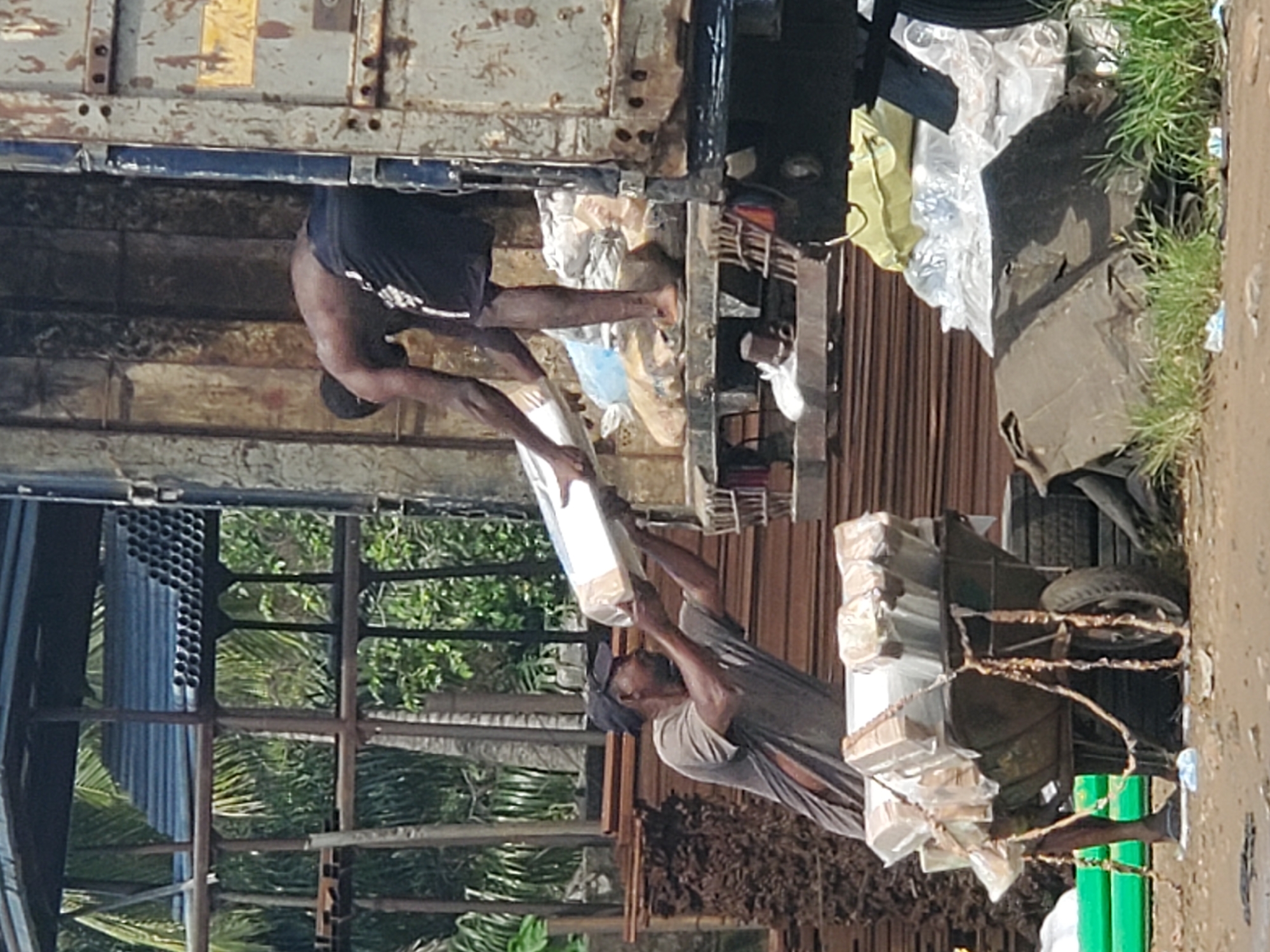 In Fegge, Nigeria, a man in a gray shirt and cap loads packages from a truck bed onto a cart. Another man wearing black shorts is inside the truck, handing items to the man below. The packages appear to be stacked on the cart, which has one visible tire. The surroundings include metal structures, vegetation, and signs of urban infrastructure.