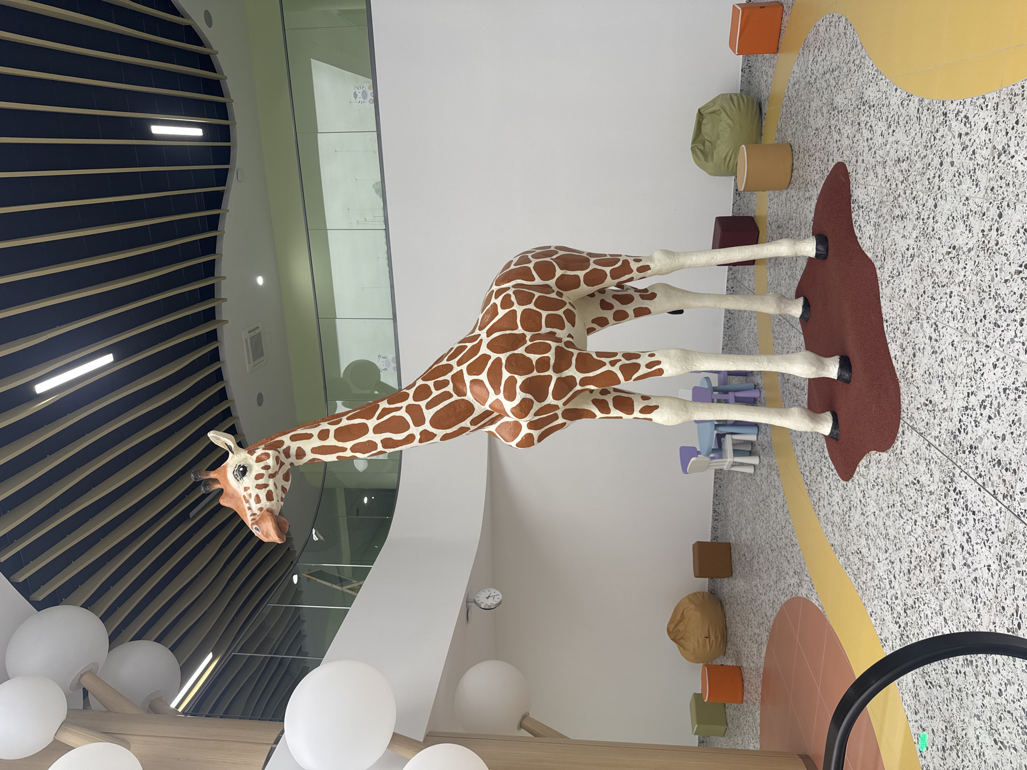 A life-sized giraffe statue stands in the center of a brightly lit, modern indoor space, possibly a children's play area or educational center. The giraffe is a realistic depiction with orange and white patterned fur and white legs. It is positioned on a dark red mat. Surrounding the statue, the floor is a speckled terrazzo with yellow and orange curvilinear patterns. In the background, there are white walls, a light-colored wall with a clock, and a railing with a green tint. Various beanbag chairs and small cube-shaped seats in green, orange, brown, and burgundy are scattered around the area. A small set of child-sized tables and chairs, colored blue and purple, is visible to the left of the giraffe's legs. The ceiling features a distinctive design with dark panels and wooden slats, interspersed with lights. Large white spherical decorations hang from a wooden structure resembling a tree on the left side of the frame. The environment appears clean, well-maintained, and designed for a young audience. The image does not depict any people or specific identifiable landmarks of Burgas, Bulgaria.