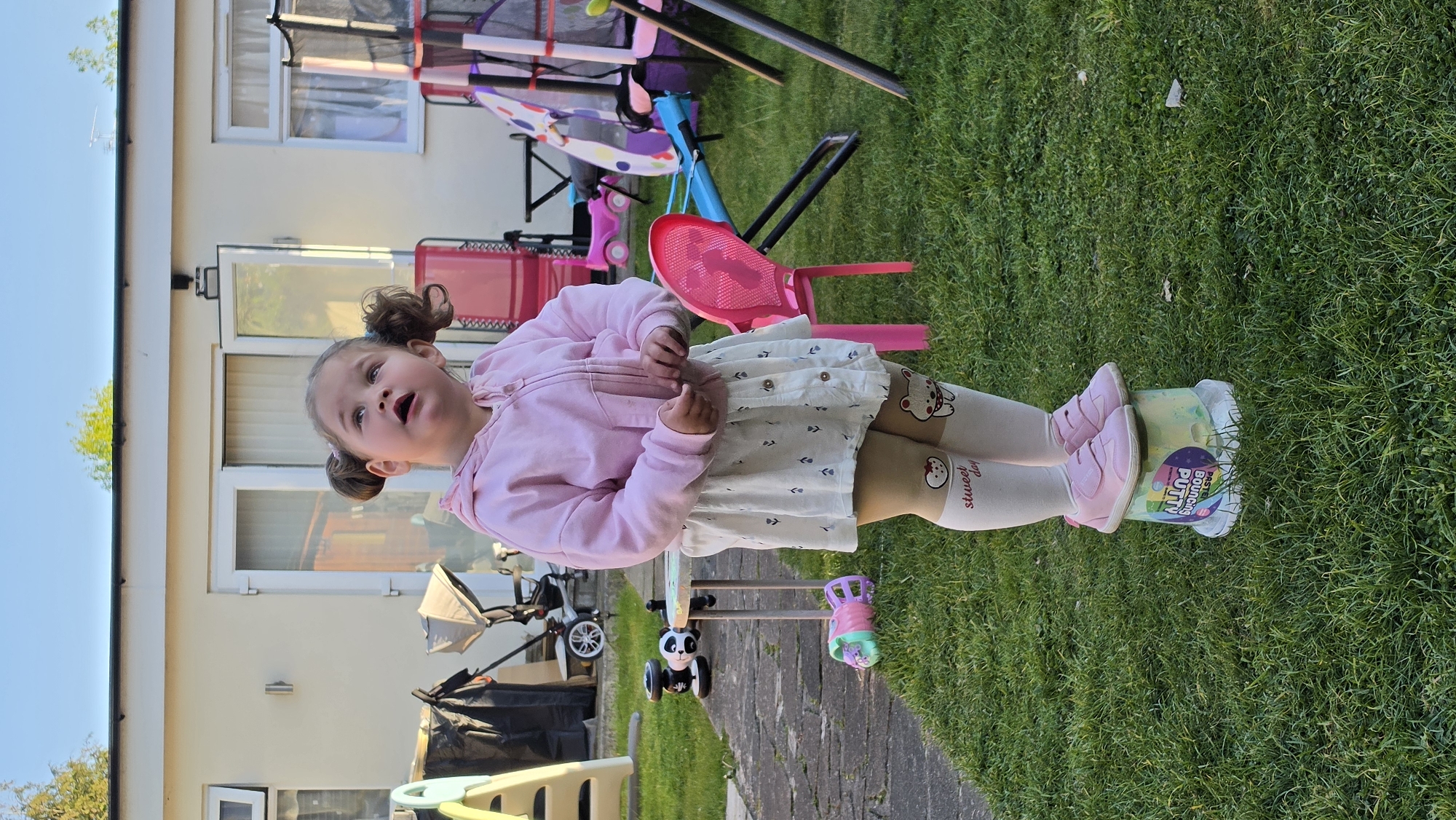 A young girl is standing in a backyard with her feet propped on a container of "Bouncing Putty." She is wearing a light pink zip-up jacket, a white skirt, and light beige leggings with cartoon bear and "sweet day" designs. She has pigtails held with pink elastics. The backyard features a green lawn, a brick path, and various children's toys including a trampoline, a pink chair, and a stroller. A white house with windows and a sliding glass door is in the background. The location is likely a residential area, and there is no information to confirm this is in the city of London, United Kingdom.