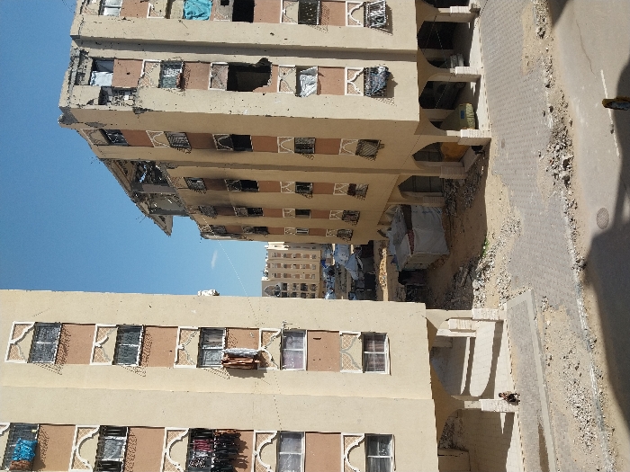 The image displays a damaged urban residential environment in Khan Yunis, Palestinian Territory, under a clear blue sky. In the foreground, two multi-story, light-colored residential buildings stand. The building on the left appears structurally sound; its facade features patterned window frames, and various pieces of clothing are hanging from several windows. At its base, a single individual is seated on a paved surface under an archway.
The building on the right exhibits substantial damage. Its uppermost floors and roof are partially collapsed and exposed, with rebar visible. The facade shows numerous impact points, broken windows, and dark scorch marks, particularly around openings, indicating fire damage. Blue tarps are visible in some window openings. Under its ground-floor arches, a vehicle covered by a green tarp is partially visible.
Between these two primary structures, the ground is unpaved and covered in significant amounts of rubble and dirt. In this area, several rudimentary shelters constructed from tarpaulins and assorted materials are visible. In the background, further multi-story buildings are present. A paved road or pathway, scattered with debris, runs across the bottom foreground. A person wearing dark clothing with a head covering is partially visible walking away in the lower right corner.