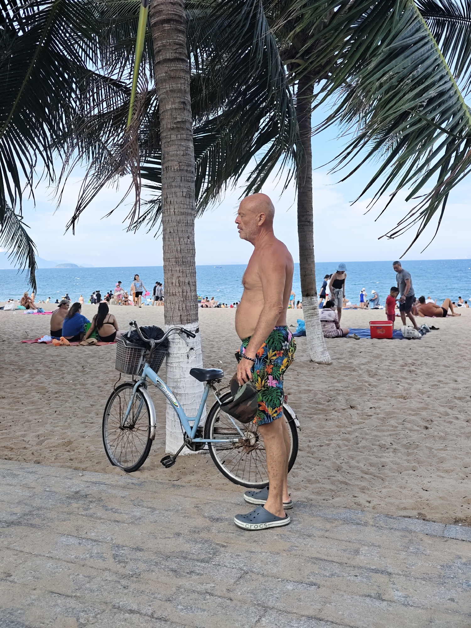 The image depicts a beach scene in Nha Trang Ward, Vietnam, under a clear sky with some clouds. In the foreground, a bald, middle-aged Caucasian man stands to the right of a light blue bicycle. He is wearing colorful, tropical-print swim trunks and dark gray Crocs. He is holding a dark helmet in his left hand. The bicycle has a basket on the front containing a black bag and a black umbrella. The bicycle is parked on a textured concrete path next to a palm tree.
The background shows a sandy beach populated with numerous people. Some are seated or lying on towels, while others are standing or walking. The ocean is visible behind the beach, with small waves and a distant shoreline. Several more palm trees are scattered across the beach. The atmosphere appears relaxed and leisurely, typical of a beach setting.