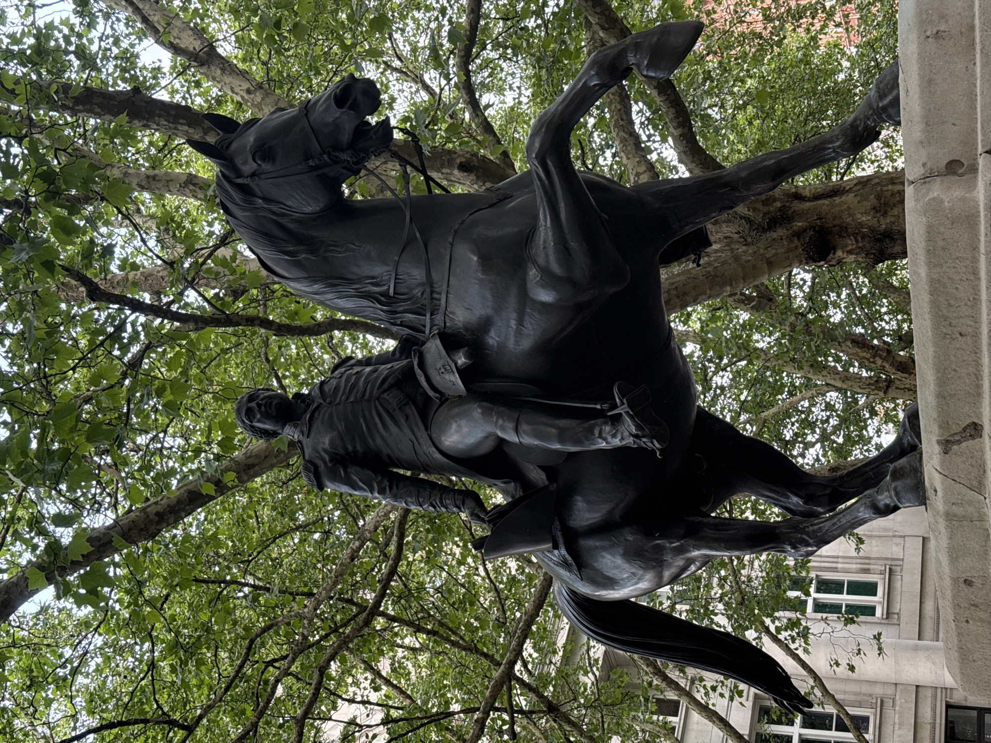 The image depicts a bronze equestrian statue of the Duke of Wellington in London, United Kingdom. The statue shows the Duke atop a rearing horse. The statue is located near Hyde Park Corner in London and is surrounded by trees with green leaves. A building can be seen in the background. The statue is mounted on a large stone plinth.
