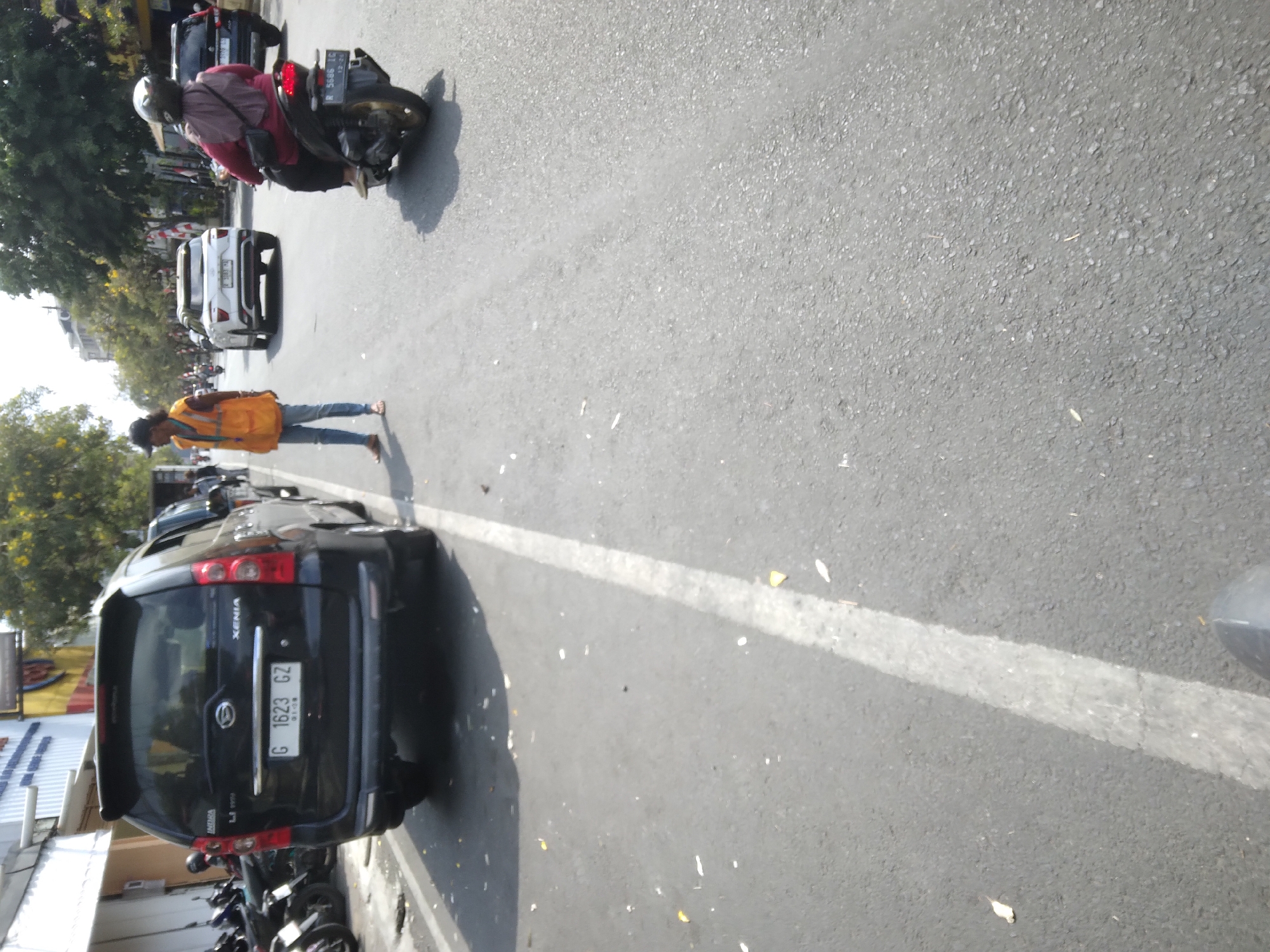 A man in an orange vest stands on a road in Purwokerto, Indonesia, looking to his right. He is positioned between a black SUV on the left and a white SUV on the right, both facing away from the camera. Behind the man, a person on a scooter with a red top and black helmet rides away from the camera. The road is paved and has a white line marking a lane. The environment appears to be a sunny day in an urban area, with trees and buildings visible in the background. Debris is scattered on the road.
