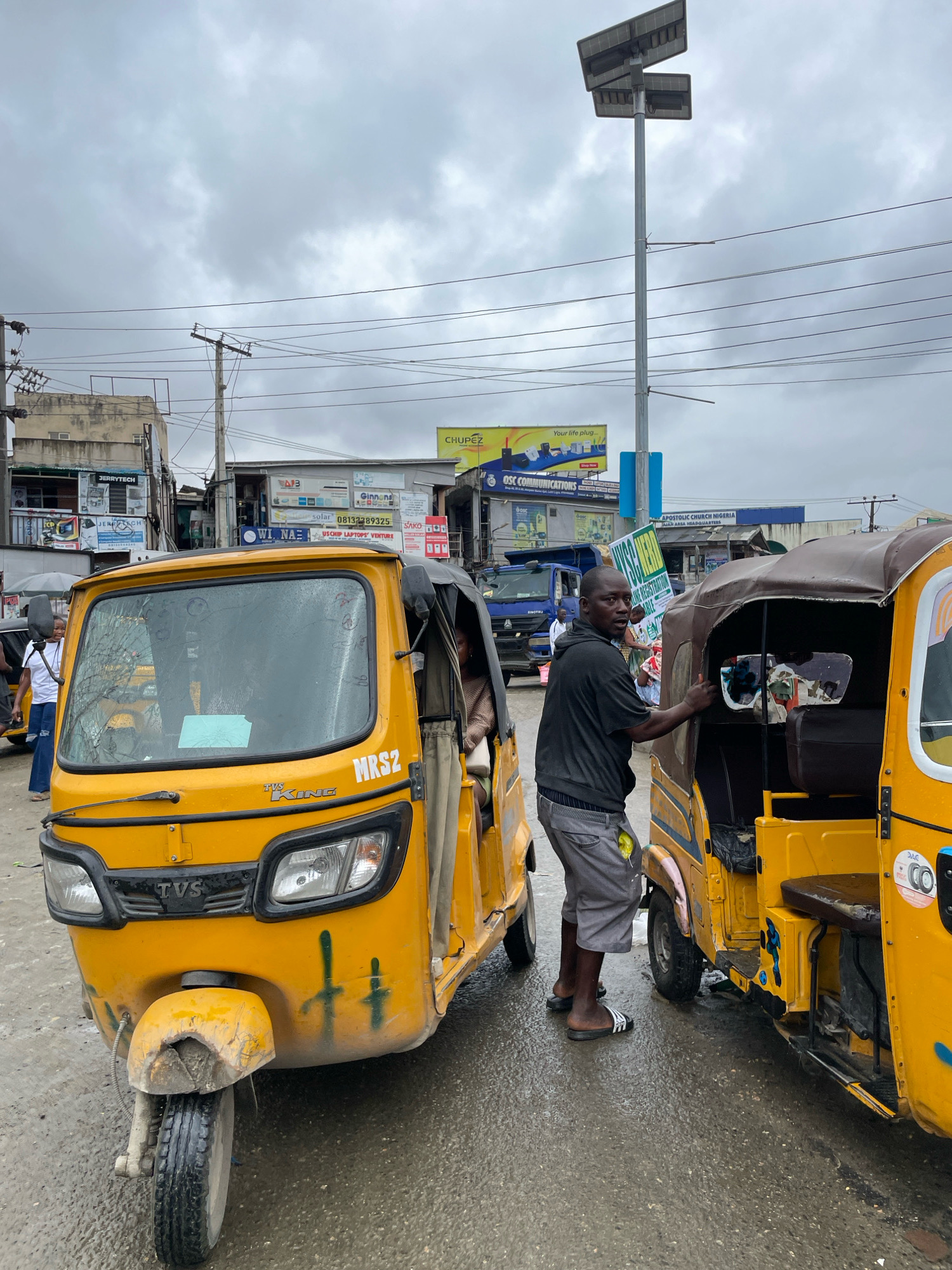 The image depicts a street scene in Ajah, Nigeria, under a cloudy sky. Two yellow auto-rickshaws, known locally as "keke napep," are prominently featured. The one on the left is a TVS KING model with a cracked windshield, displaying "MRS2" on its front panel. The rickshaw on the right has a brown canopy and is partially obscured by a man standing next to it. This man, wearing a dark hooded sweatshirt and gray shorts, appears to be interacting with the rickshaw driver or passenger. Another person, a woman in a light-colored top and blue jeans, is visible in the background near the left rickshaw. Various buildings with signage indicating businesses like "JERRYTECH," "CHUPÉZ," "OSC COMMUNICATIONS," and "APOSTOLIC CHURCH NIGERIA" line the street, suggesting a commercial area. Power lines and a solar-powered street light are visible overhead. The ground is wet, indicating recent rain. The overall impression is that of a busy, somewhat weathered urban environment.