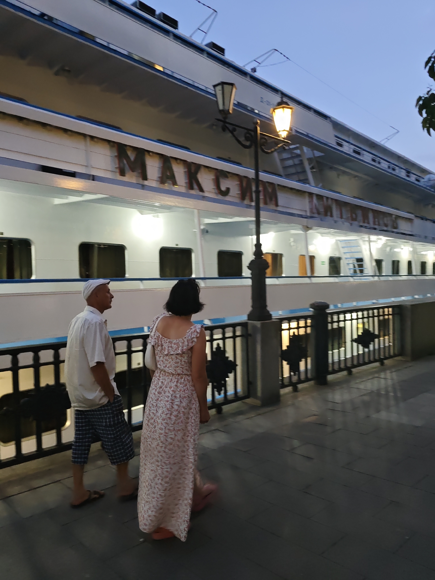 The image captures a view of the steamer "Maksym Gorky" docked in Rostov-on-Don, Russia, alongside the Don River. Two individuals, a man and a woman, are walking away from the camera on a paved walkway. The man, wearing a white short-sleeved shirt, a white cap, and plaid shorts, walks with his hands in his pockets. The woman, in a floral-patterned dress and carrying a white purse, walks slightly ahead of him. A decorative black metal fence separates the walkway from the river. In the background, the large white passenger ship is illuminated, with visible windows and signage that reads "МАКСИМ ГОРЬКИЙ" in Cyrillic. A lamppost with two lights is positioned between the couple and the ship, casting a warm glow on the scene, which appears to be set during dusk or early evening.