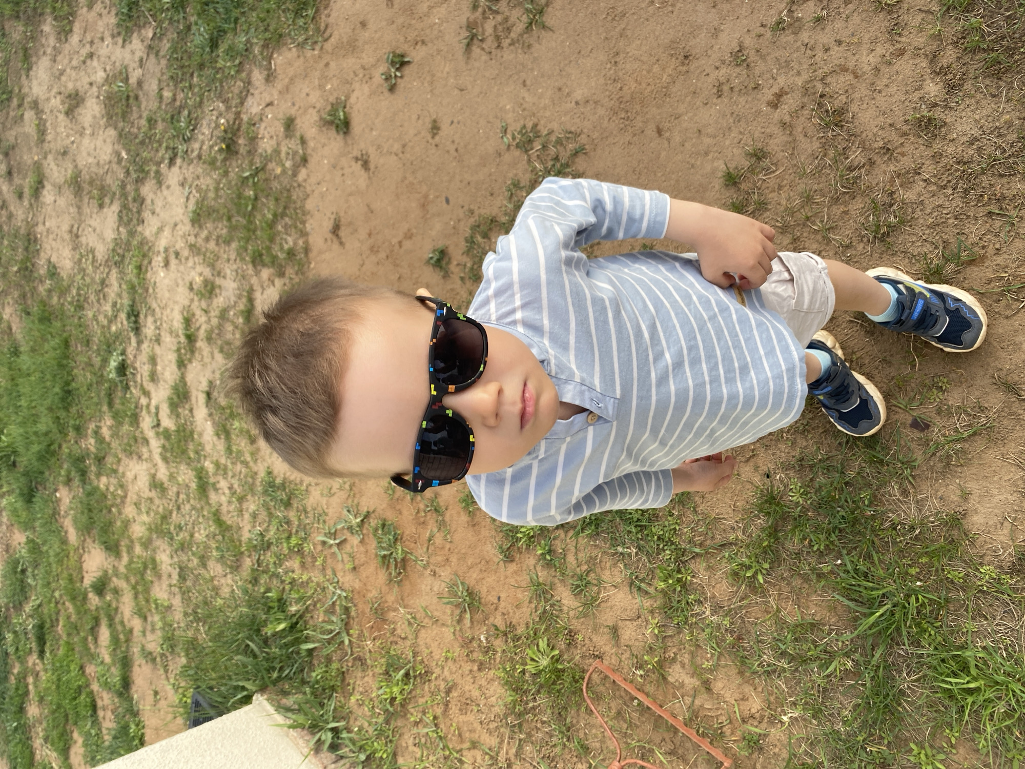 A young boy stands outdoors on a patch of mixed grass and dirt. He is wearing sunglasses, a light blue and white striped long-sleeved shirt, beige shorts, and blue sneakers with white trim. The ground consists of bare earth interspersed with patches of green grass and weeds. The location appears to be in Chaikovsky, Russia.