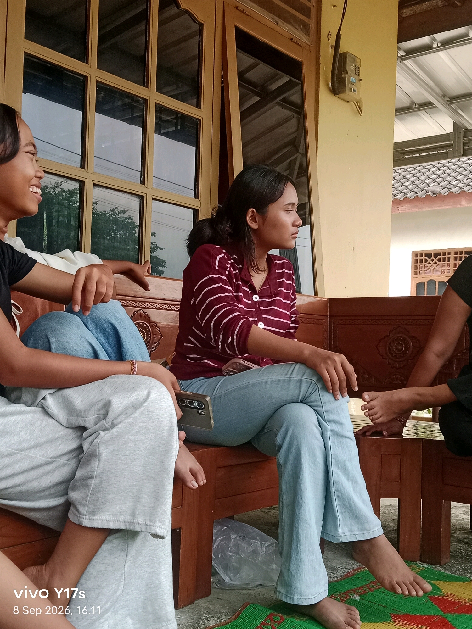 Three individuals are seated on a wooden bench in what appears to be an outdoor, covered area. The person on the left, a child, is wearing a black t-shirt and grey pants, smiling broadly and looking to their left. To the right of the child, a young woman is wearing a maroon and white striped shirt and light blue jeans, looking to her right with a neutral expression. Her legs are crossed. To the far right, another person's feet and lower legs are visible, wearing dark clothing. They are also seated on the bench. The background includes a wooden window frame with multiple panes, a light yellow wall, and a corrugated metal roof. An electrical meter is mounted on the wall. A woven mat is on the ground in front of the bench. The location is identified as Rama Dewa, Indonesia. The timestamp on the image indicates it was taken on September 8, 2026, at 4:11 PM.