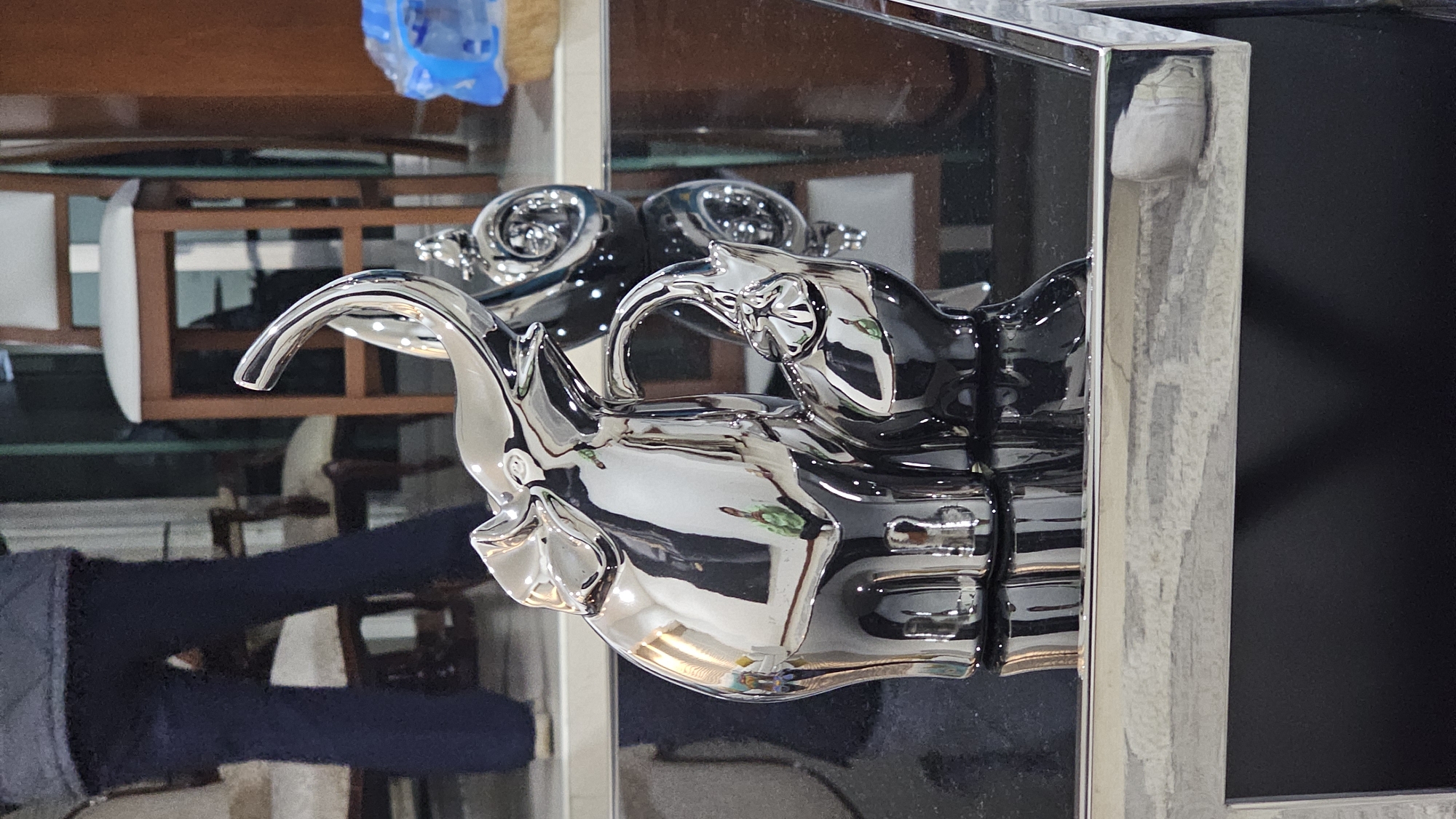 The image shows a silver-colored metal elephant figurine mounted on a mirrored surface. The figurine is positioned against a dark background, possibly a television screen or a similarly dark surface. The reflective surface of the mirror creates multiple reflections of the figurine and the surrounding environment. The reflections suggest that the figurine is displayed in a setting with other furniture and objects, visible in a blurred manner. The location of San Bernardino Tlaxcalancingo, Mexico, is not evident from the visual content of the image.