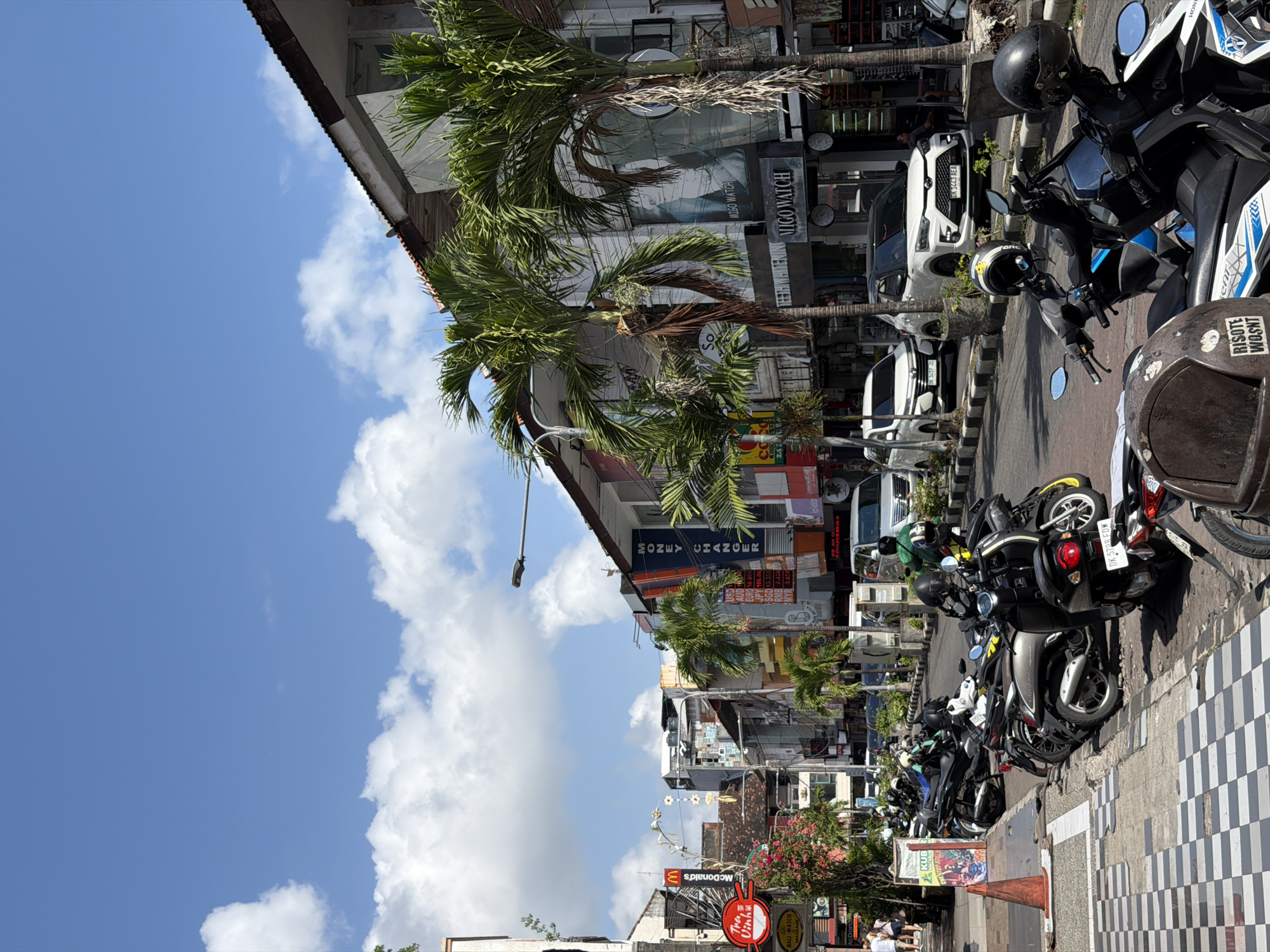 The image depicts a street scene in Kuta, Indonesia, on a bright, sunny day with scattered clouds. The perspective is from a slightly elevated, tilted angle, giving a dynamic view of the bustling street.
The main subjects are a row of parked motorcycles and scooters lining the sidewalk. Several people are visible, some walking along the street and others sitting near a building. The scene is dominated by commercial buildings, many with signs advertising shops, restaurants, and services. Notable text includes "McDonald's," "MONEY CHANGER," "MIGO WATCH," and "BALI WATCH." The presence of palm trees adds to the tropical ambiance of the location.
The overall impression is one of a busy urban environment, likely during daytime, given the strong sunlight and clear sky. The parked vehicles suggest a popular area where people arrive by scooter. The tiled pattern on the sidewalk and the architecture of the buildings are characteristic of streetscapes in this region. The image captures the vibrant and commercial atmosphere of a tourist-oriented area in Kuta.