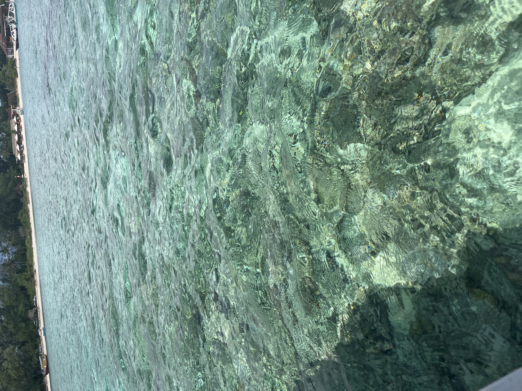 The image captures a clear, shallow body of water characterized by its vibrant light turquoise color and rippling surface. The transparency of the water allows the sandy and rocky seabed, featuring various textures and darker patches of what might be coral or marine growth, to be distinctly visible beneath the sunlight.
To the left, a narrow strip of white sandy beach extends along the water's edge. Beyond the beach, a dense line of lush green tropical vegetation, consisting of trees and bushes, forms the backdrop. Along the beachfront, several small structures, possibly resort buildings or huts, are visible, accompanied by white beach chairs and umbrellas spread across the sand. A small boat or part of a boat can also be faintly discerned near the shoreline.
The scene depicts a peaceful tropical setting under bright daylight, with sunlight creating luminous patterns on the water's surface and the visible seabed. No people or specific activities are observable, and there is no visible text.
