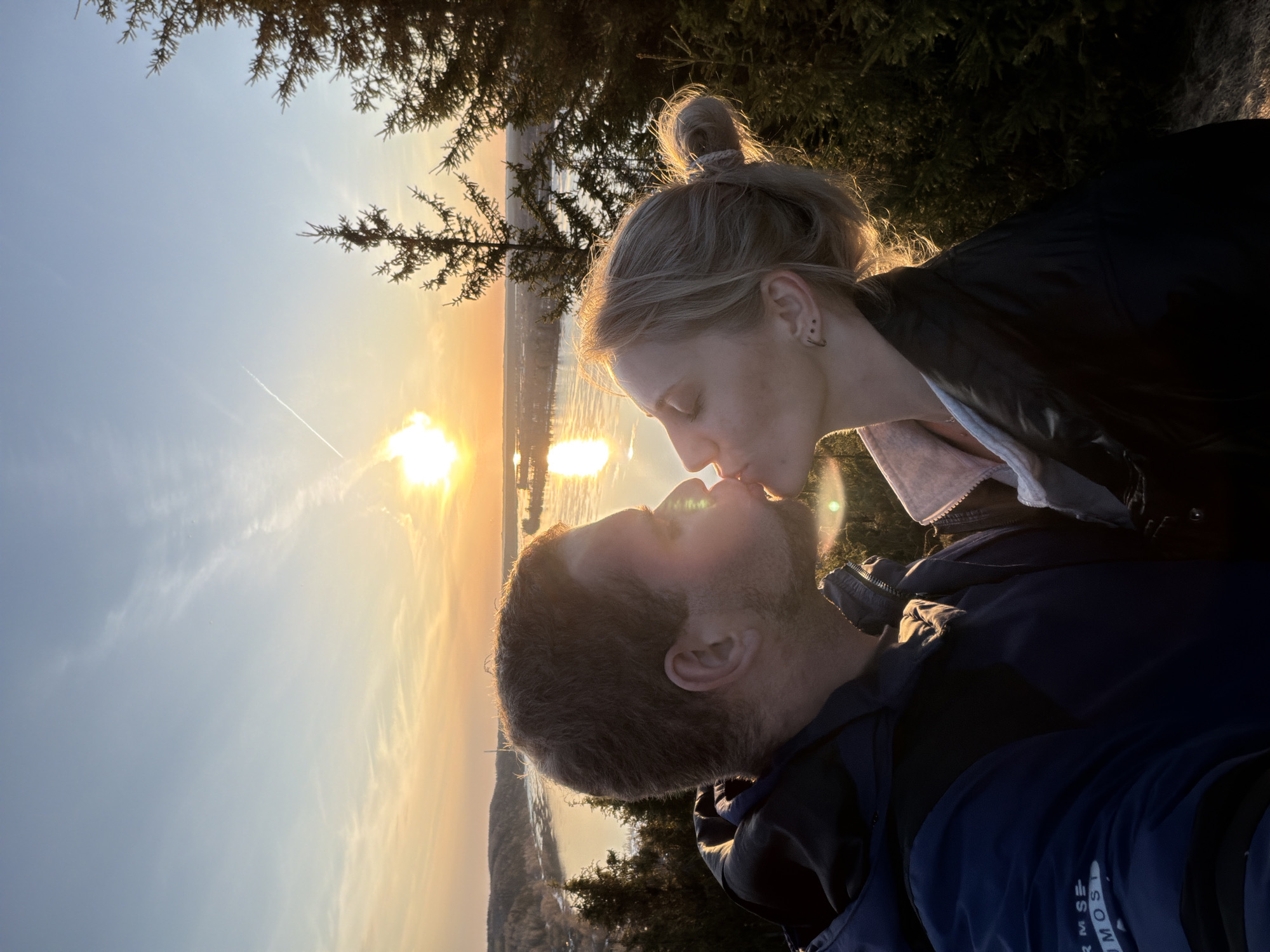 A medium shot captures a man and a woman kissing at sunset, likely in Ольховка, Russia, given the location provided. The woman has fair skin and her blonde hair is styled in a bun. She is wearing a dark jacket over a light-colored garment, possibly a blouse or shirt. The man has short brown hair and a beard; he is wearing a blue jacket. The setting sun is visible behind them, casting a golden light on the scene. The background includes a body of water and vegetation, suggesting they are in a park or on a hill overlooking a lake or river.