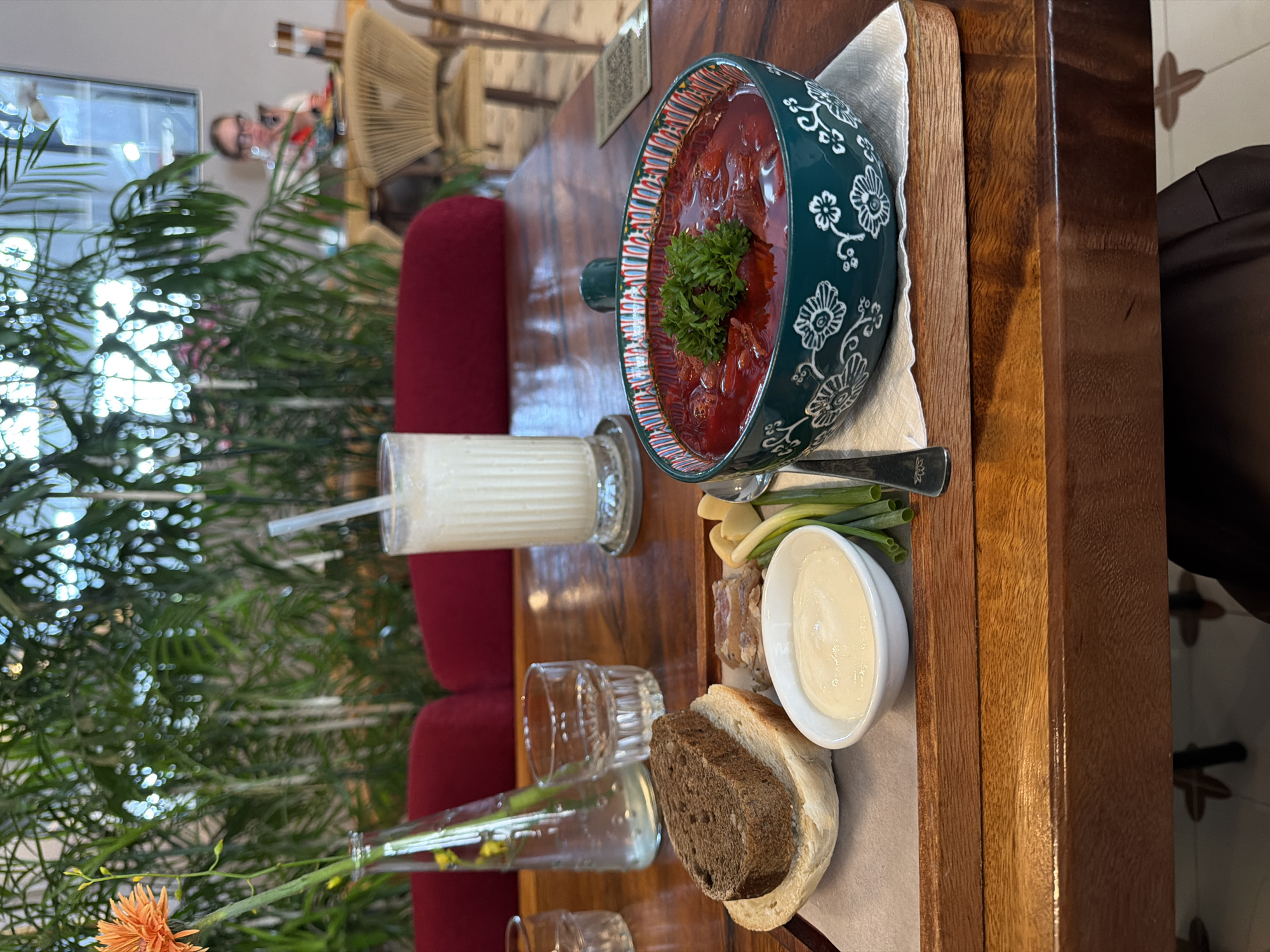 A wooden table in what appears to be a restaurant or cafe in Da Nang, Vietnam, holds a meal and drinks. In the foreground, a wooden tray contains a slice of dark bread on top of a white bread roll, a small white bowl filled with a creamy white sauce, some sliced meat, a few green onions, and a spoon and fork. Beside the tray, a dark green bowl with a white floral pattern is filled with a vibrant red soup, garnished with green herbs. A tall glass of a light-colored beverage with a straw stands next to the soup. In the background, a vase with an orange flower and greenery is visible. Beyond that, red chairs and other tables are set up, with a person seated at a table in the far background, looking at a phone. The overall ambiance suggests a relaxed dining setting, possibly with an indoor garden element due to the abundance of plants.