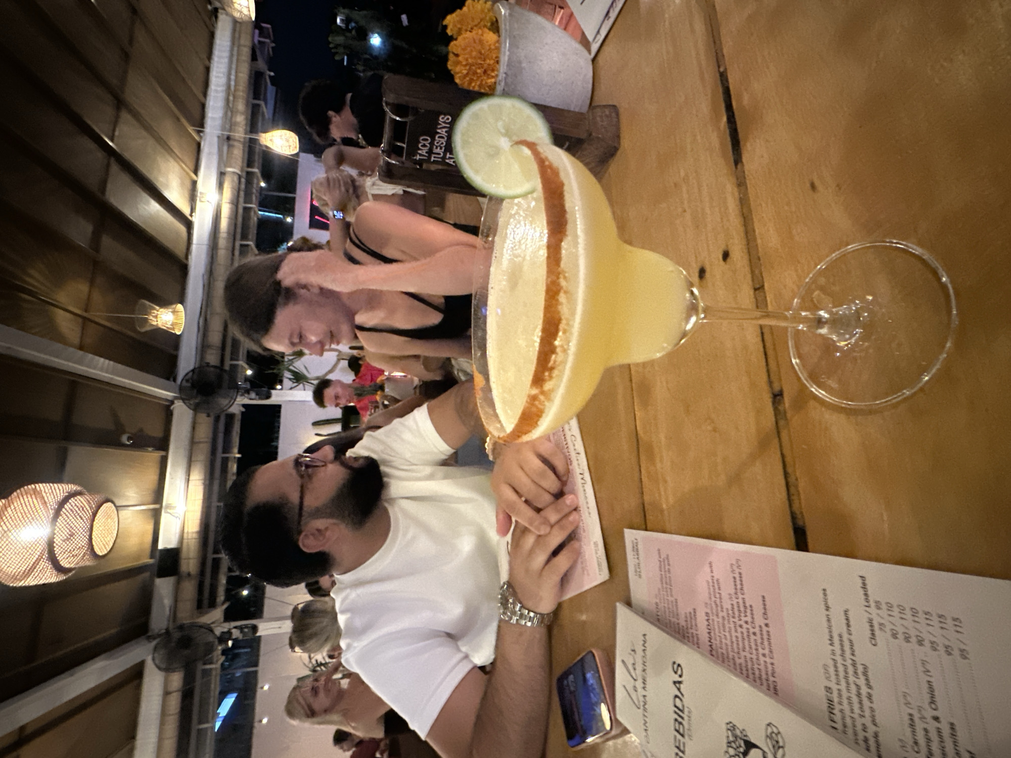 A couple is seated at a wooden table in a restaurant, presumably in North Kuta, Indonesia, judging by the menu from "Lola's Cantina Mexicana." A margarita, garnished with lime, is prominently placed on the table. The man is wearing a white t-shirt and a watch, and is looking at the woman. She's wearing a black tank top. A menu is also present. In the background, other patrons and restaurant décor, including hanging lights, are visible. There is a small sign that reads "TACO TUESDAYS AT."
