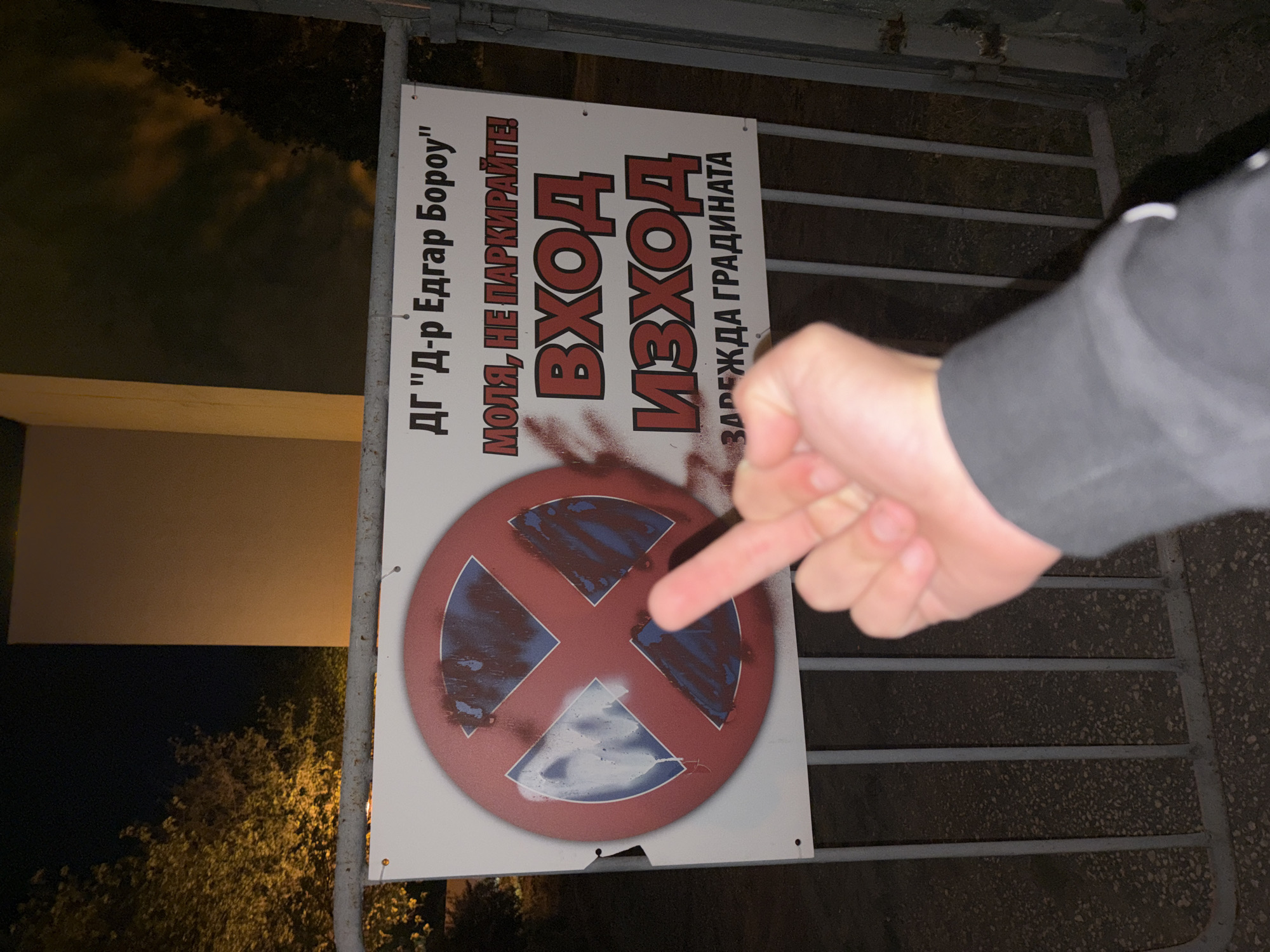 The image captures a vandalized "No Parking" sign, likely in Plovdiv, Bulgaria, though this is not explicitly stated. The sign reads "ДГ 'Д-р Едгар Бороу' МОЛЯ, НЕ ПАРКИРАЙТЕ! ВХОД ИЗХОД ЗАРЕЖДА ГРАДИНАТА", indicating it's associated with a Dr. Edgar Borou kindergarten. The sign's circular "no parking" symbol has been obscured with what appears to be blue and brown spray paint. A person's hand, specifically the middle finger, is pointed at the vandalized sign. The sign is affixed to a metal gate or fence. The environment is dark, suggesting nighttime, with foliage visible in the background and a wall in the periphery.