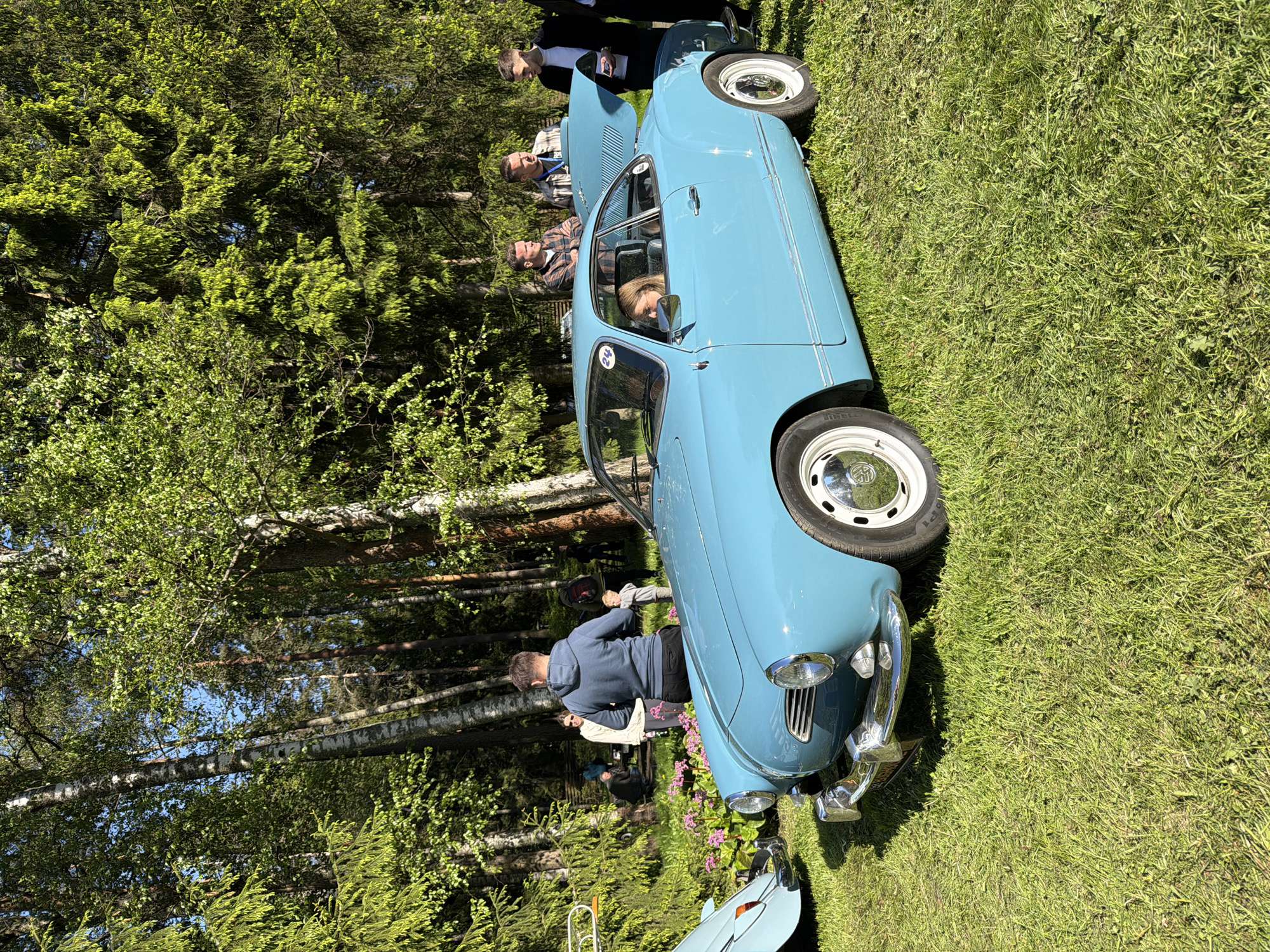 The image depicts a light blue vintage car parked on a grassy field, likely in Repino, Russia. The car has its hood open, revealing part of the engine. Several people are standing around the car, some examining it, others conversing. The environment includes trees and sunlight, suggesting an outdoor car show or gathering. There are other cars in the background, with the front of one just visible.