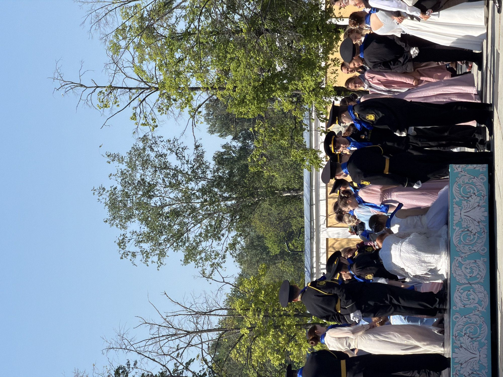 A formal outdoor event takes place in Kronstadt, Russia. It features a group of young people in historical attire, some in military-style uniforms and others in long gowns. They are aligned on a decorative platform. The sky is blue, and trees frame the scene.