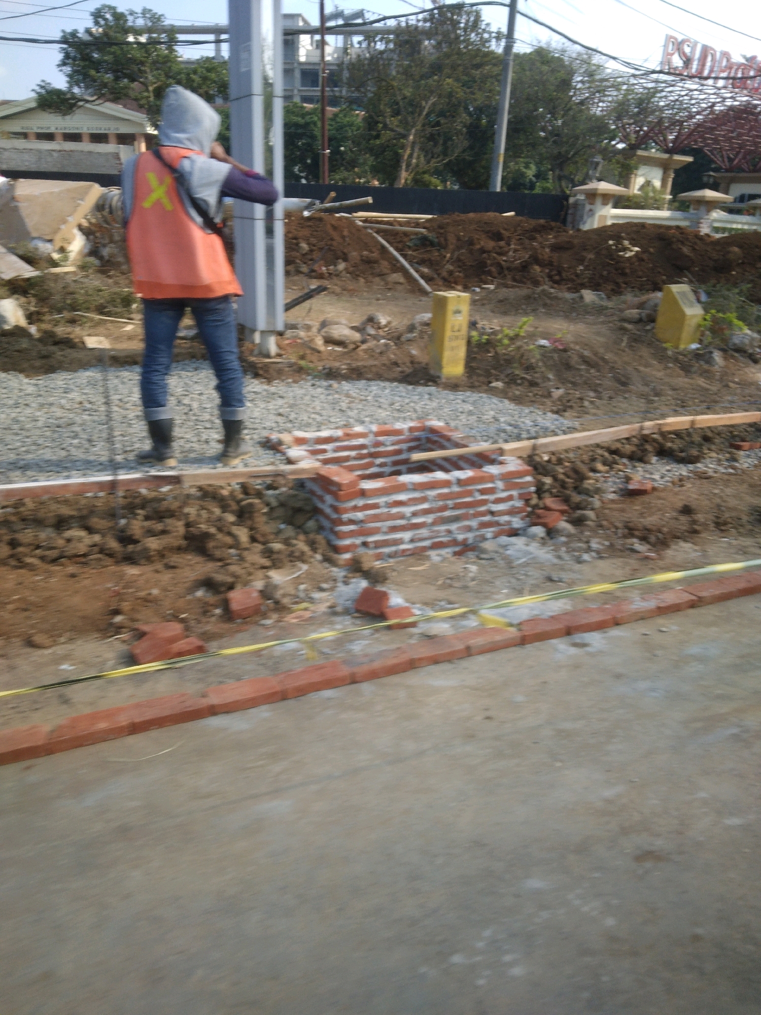 A construction worker, wearing a grey hooded jacket and an orange reflective vest with a yellow "X" on the back, stands in a construction area. They are also wearing blue jeans and black boots. The worker is facing away from the camera and appears to be holding a dark object to their ear, possibly a phone. The surrounding environment is a dirt and gravel construction site with piles of dirt, bricks, and construction materials. A partially constructed brick structure is visible. In the background, there are buildings, trees, and utility poles, suggesting an urban or suburban setting. The overall scene depicts ongoing construction work.