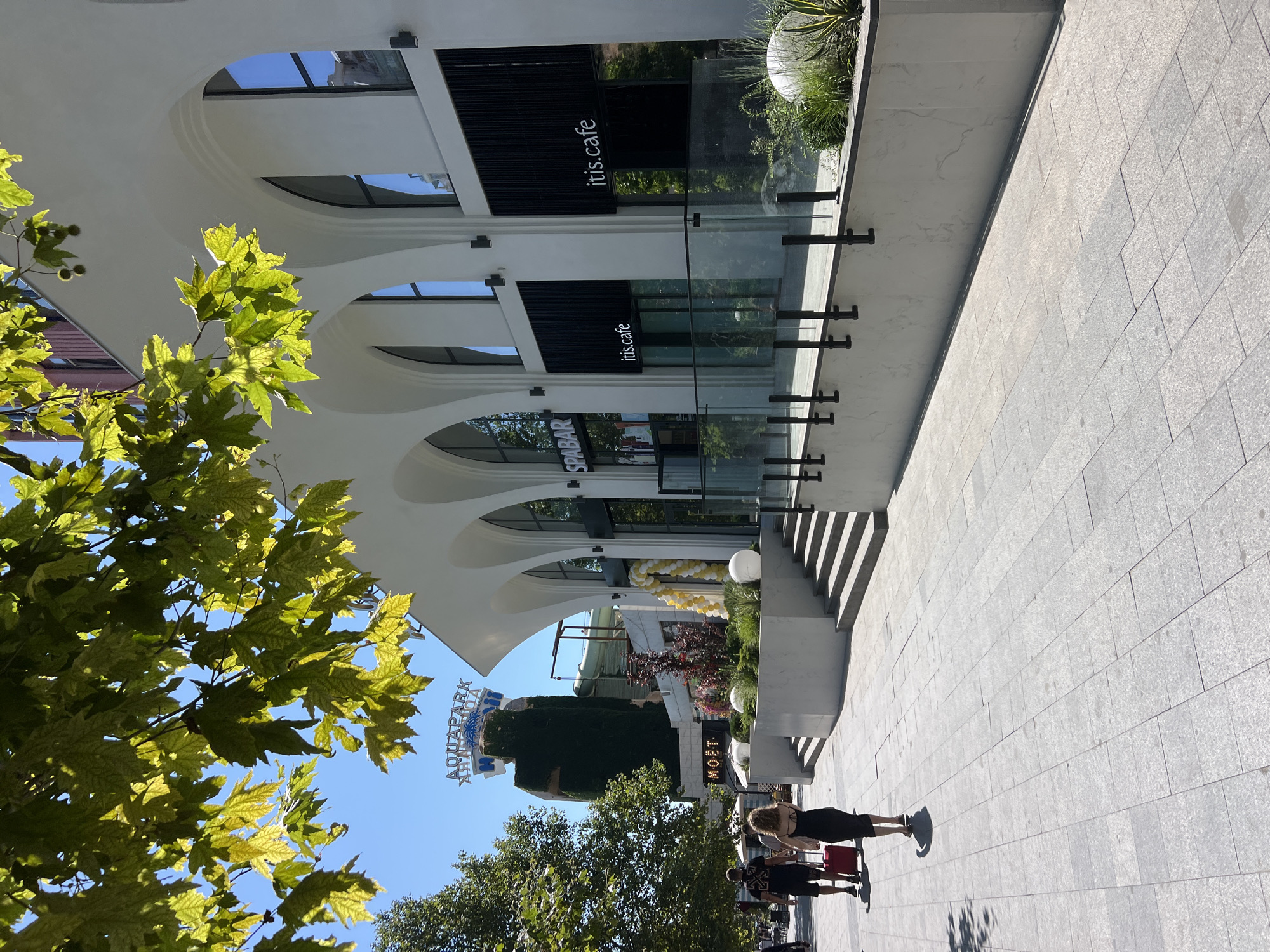 In an outdoor urban setting, a modern white building with arched windows is visible on a sunny day. The building's facade features several large, arched windows and dark-colored awnings or signage that reads "SPABAR" on one section and "itis.cafe" on two others. In front of the building, a grey tiled walkway leads away from the viewer. Two individuals are walking on the walkway; a man in a black t-shirt and shorts is walking ahead, and a woman in a black dress is walking behind him, pulling a red suitcase. To the left of the walkway, trees and a large, vine-covered structure topped with signage reading "AQUAPARK" are present. Lush greenery and large white spherical decorations are visible on a raised planter area in front of the building. The sky is clear and blue.