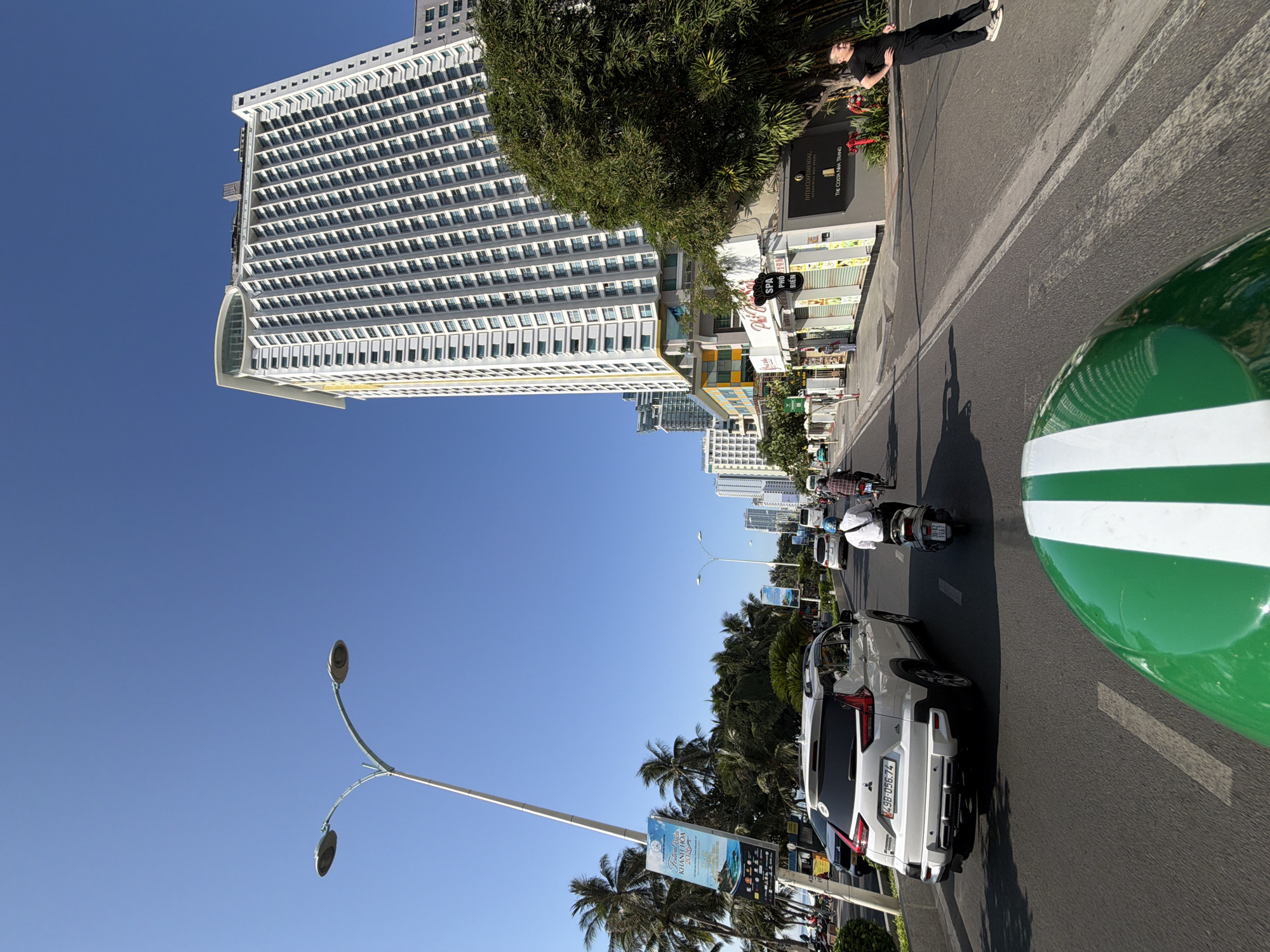 The image depicts a sunny day on a busy street in Nha Trang, Vietnam. In the foreground, a green and white object, possibly a helmet, suggests the perspective of a motorcyclist. The street is paved and lined with vehicles, including a white Mitsubishi Xpander SUV with a license plate reading "498-056.74" and several motorbikes with riders. A man in a black shirt and pants stands on the sidewalk on the right, facing away from the camera with his hands on his hips.
Dominating the skyline are several tall, modern buildings. The most prominent is a white, multi-story hotel with a distinctive curved glass structure at its peak. Adjacent to it, other buildings of varying heights line the street. On the left side of the road, palm trees are visible, and a lamppost with two lights stands tall, adorned with several vertical banners. One banner advertises "Festival Nha Trang Khanh Hoa 2023." Further down the street, another banner with similar content is also visible. Shops and storefronts are present along the sidewalk, including one with the sign "SPA PHU BIEN." The overall environment suggests a vibrant urban setting, likely a tourist area, with clear blue skies overhead.
