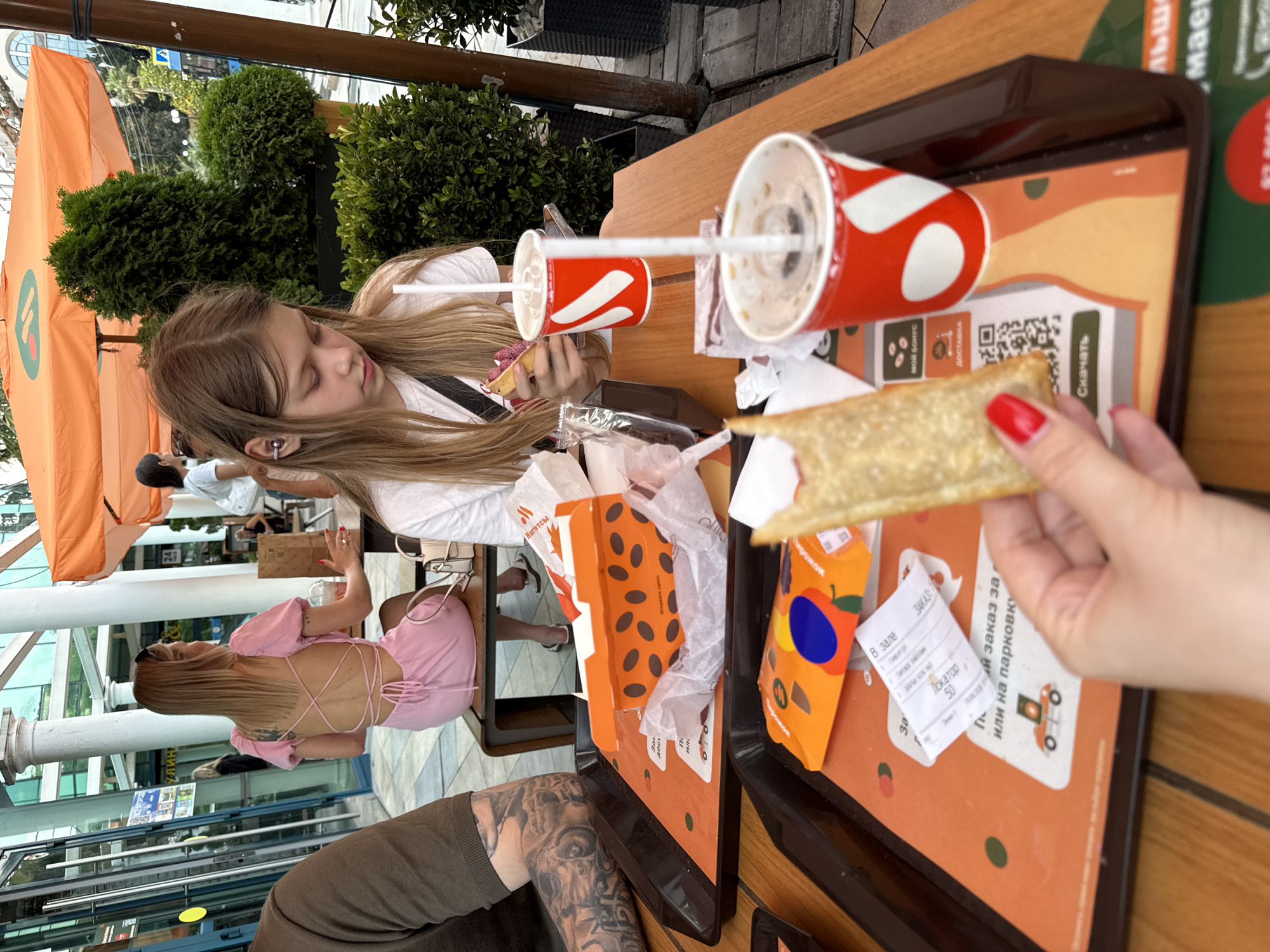 This image depicts an outdoor dining scene at a fast-food establishment in Sochi, Russia. The foreground features two black plastic trays filled with fast food and drinks. One tray holds a red and white branded cup with a straw, a paper bag, and an orange box with dark brown oval patterns, partially opened. The second tray, closer to the viewer, contains another branded cup with a straw, an orange package with illustrations of fruit and a blue circle, and a receipt. A hand with bright red nail polish is holding a partially eaten food item, possibly a wrap or pastry, over this tray.
In the middle ground, a young girl with long blonde hair and earbuds is seated at a wooden table, looking down at her food and drink. She is wearing a white t-shirt. Behind her, a woman with blonde hair tied back with a black headband is seated, wearing a pink backless dress with crisscross straps. She is also at a table, with a drink in front of her.
The background reveals more of the establishment, including an orange umbrella providing shade and a glimpse of a building with large glass windows. Lush green bushes and trees are visible, suggesting an outdoor or semi-outdoor seating area. The overall setting is casual and typical of a fast-food restaurant with outdoor seating in a warm climate. The presence of Cyrillic text on the packaging and receipt indicates the location is Russia, and the mention of "Sochi" in the prompt suggests this is the specific city.