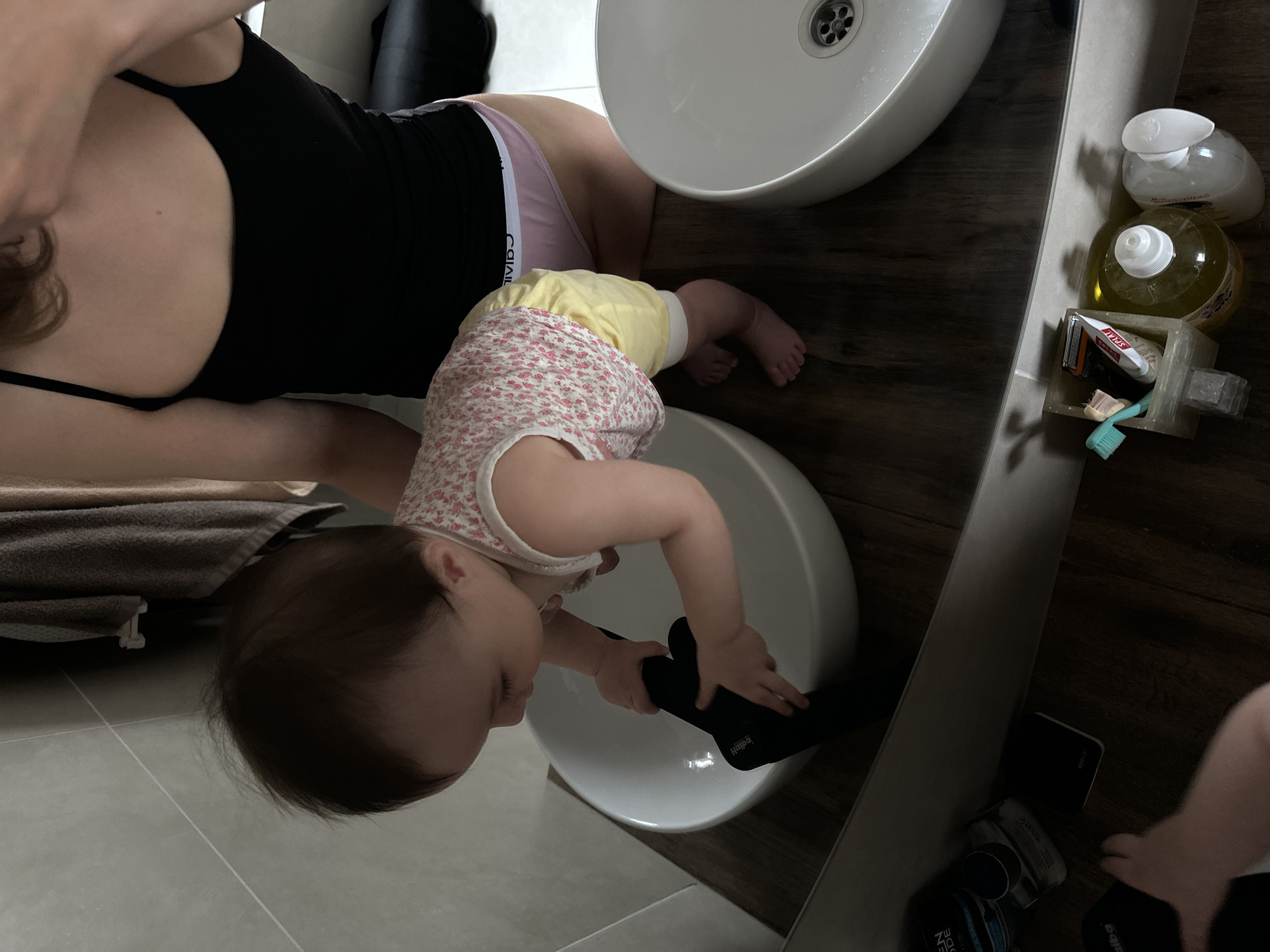A baby in a floral and yellow outfit is playing with a black object inside a white sink. An adult is nearby, partially visible wearing a black top and Calvin Klein underwear. Two sinks are present in a bathroom setting with a wood-look countertop. Toiletries are near the sink, and a portion of the tile floor is visible. The exact location is Подшивалово, Russia.