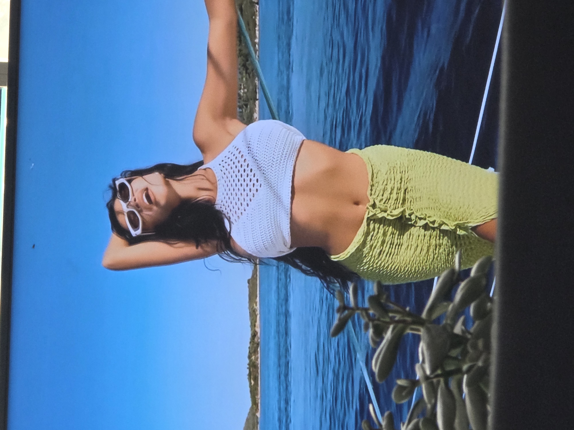 A person with dark hair, wearing white cat-eye sunglasses, a white crochet halter top, and a ruched lime green skirt, is lying on their back with their head tilted back and arms behind their head. They are outdoors, with a bright blue sky and a body of water in the background. The water appears to be wavy and a deep blue color, possibly the sea. There's a hint of a green, rocky shoreline on the left side of the image. In the foreground, there's a blurry section of dark green foliage. The lighting suggests it's daytime, possibly sunny given the bright blue sky. The overall mood seems relaxed and carefree.
