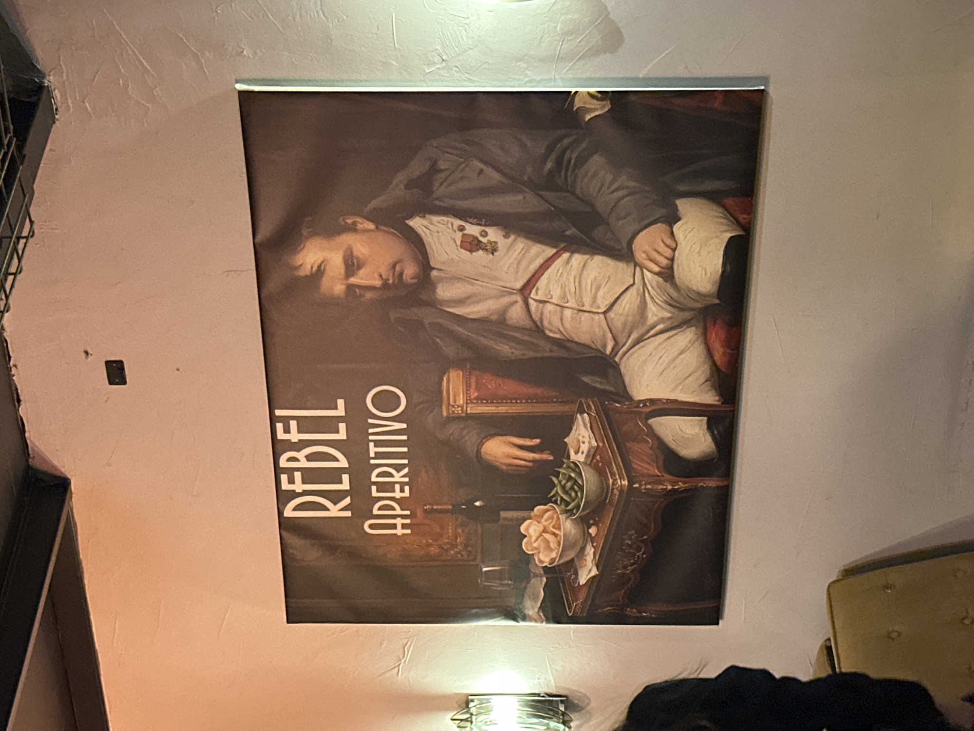The image shows a poster advertising "REBEL APERITIVO" hanging on a textured, light-colored wall. The poster depicts a reclining figure resembling Napoleon Bonaparte, dressed in a white uniform and a dark jacket, with a table displaying an array of snacks and a bottle of wine in front of him. The environment appears to be an interior, possibly a bar or restaurant.
The location in Milan, Italy, is inferred from the word "APERITIVO" on the poster, aperitivo being an Italian custom of pre-dinner drinks with snacks.