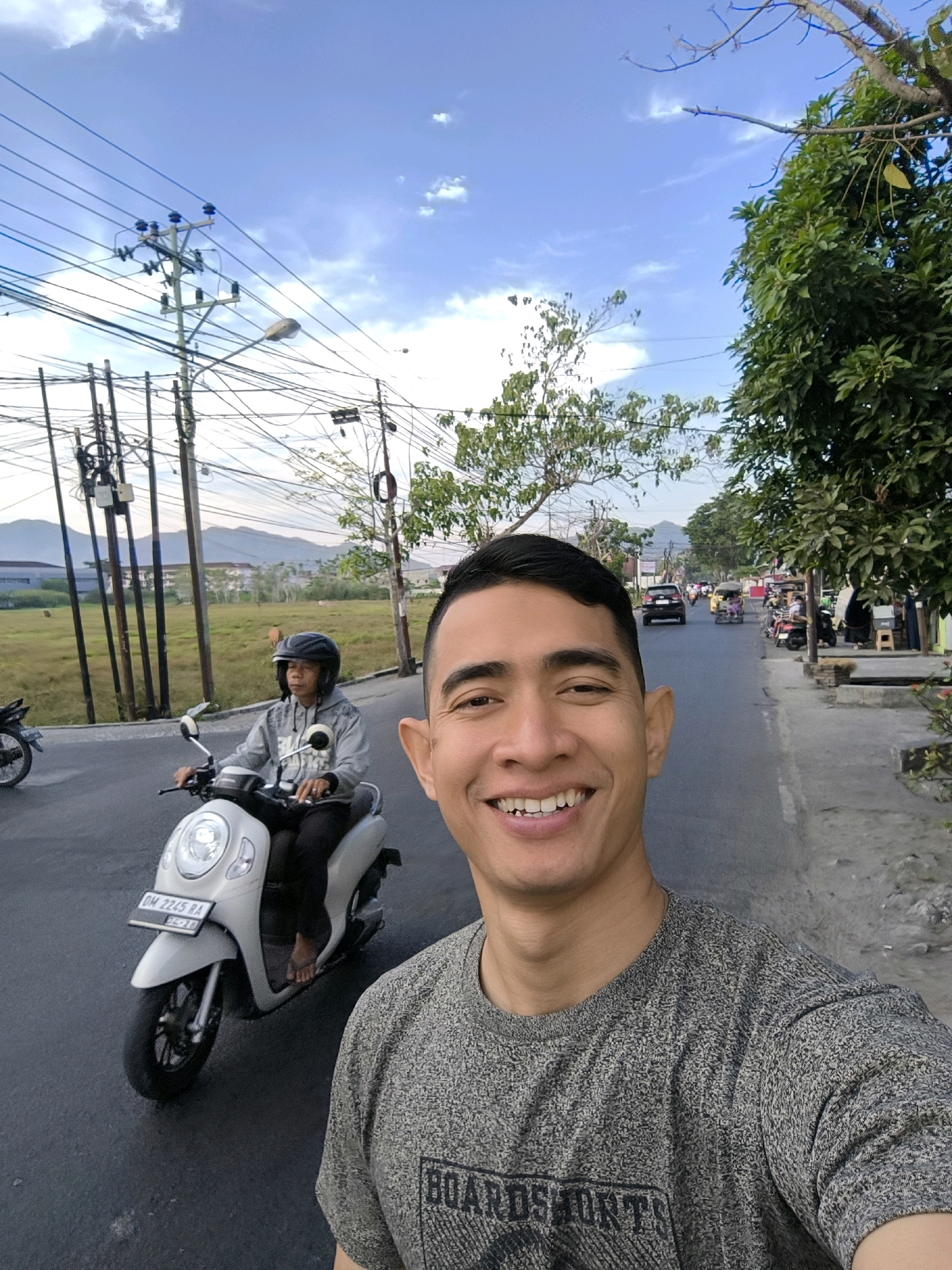 A man in a gray t-shirt is taking a selfie on a street in Gorontalo, Indonesia. He is smiling and looking at the camera. Behind him, another person is riding a white scooter and wearing a helmet. The road is paved, and there are utility poles with many wires on the left side of the street. Trees and buildings line the right side of the road. In the background, there are mountains and a cloudy blue sky.