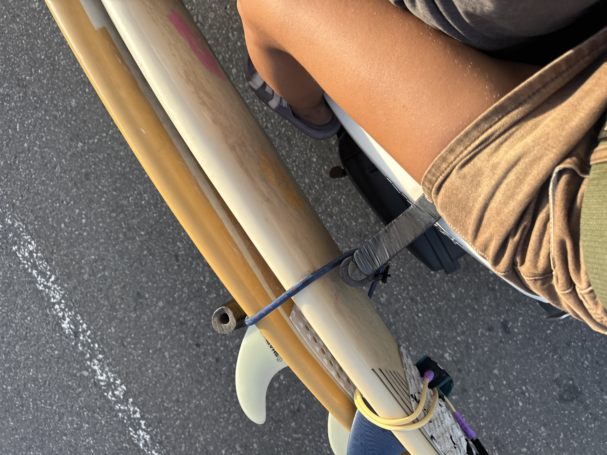 In Denuwala, Sri Lanka, three surfboards are being transported, secured to a vehicle by a blue bungee cord and straps. The surfboards are layered diagonally, with the top board appearing to be a bright yellow or orange, the middle board a lighter cream color with pink markings, and the bottom board a similar cream color with black lines. A light-colored fin with the brand "SHAPER" is visible on the middle surfboard. A person's legs, tanned and wearing brown shorts, are positioned in the foreground, suggesting they are either the operator or passenger of the vehicle. The surfboards are positioned over a dark, possibly black, object, which could be part of the vehicle's rack or frame. The background consists of dark asphalt with a faint white line, indicating a road surface. The lighting suggests it is daytime, with shadows cast on the road.
