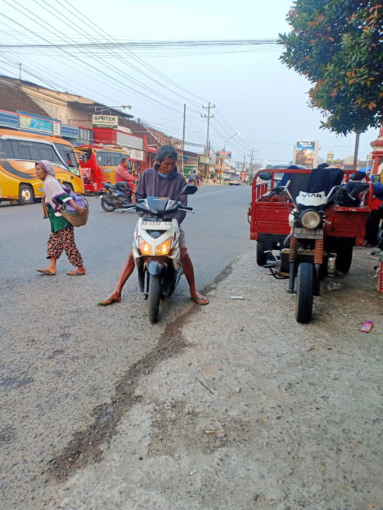 The image depicts a street scene in Mojokerto, Indonesia, likely during the early morning or late afternoon given the soft lighting. In the foreground, a man with graying hair is seated on a white and black Honda scooter, wearing a dark purple long-sleeved shirt and shorts. His legs are visible as they straddle the scooter. To the left, an elderly woman is walking, carrying a woven basket filled with items on her left shoulder. She wears a light pink headscarf, a green shirt, and patterned pants, with her feet in flip-flops. Behind the scooter, a red three-wheeled motorcycle, identified by the "VIAR" logo on its front, is parked. This vehicle has a cargo bed laden with dark-colored fabric. The street is paved with asphalt and shows signs of wear and tear, with some debris and water patches. In the background, various buildings line the street, including shops with signs like "APOTEK DHARMA SEHAT". Several vehicles, including a yellow bus, are present on the road. Overhead, a network of power lines crisscrosses the sky. A large tree with dense foliage is visible on the right side of the image. The overall impression is of a busy, functional street in a Southeast Asian urban environment.