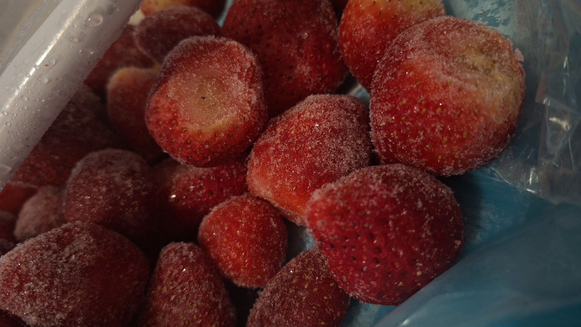 The image shows a close-up view of a bag of frozen strawberries. The strawberries are red and covered in a layer of frost. The bag is made of clear plastic and has some water droplets on its surface. A portion of a blue container is also visible, possibly the bag's resting place. No people, subjects, objects, actions, interactions or other environmental context related to the location in Забалоцкі сельскі Савет, Belarus are discernable from the image.