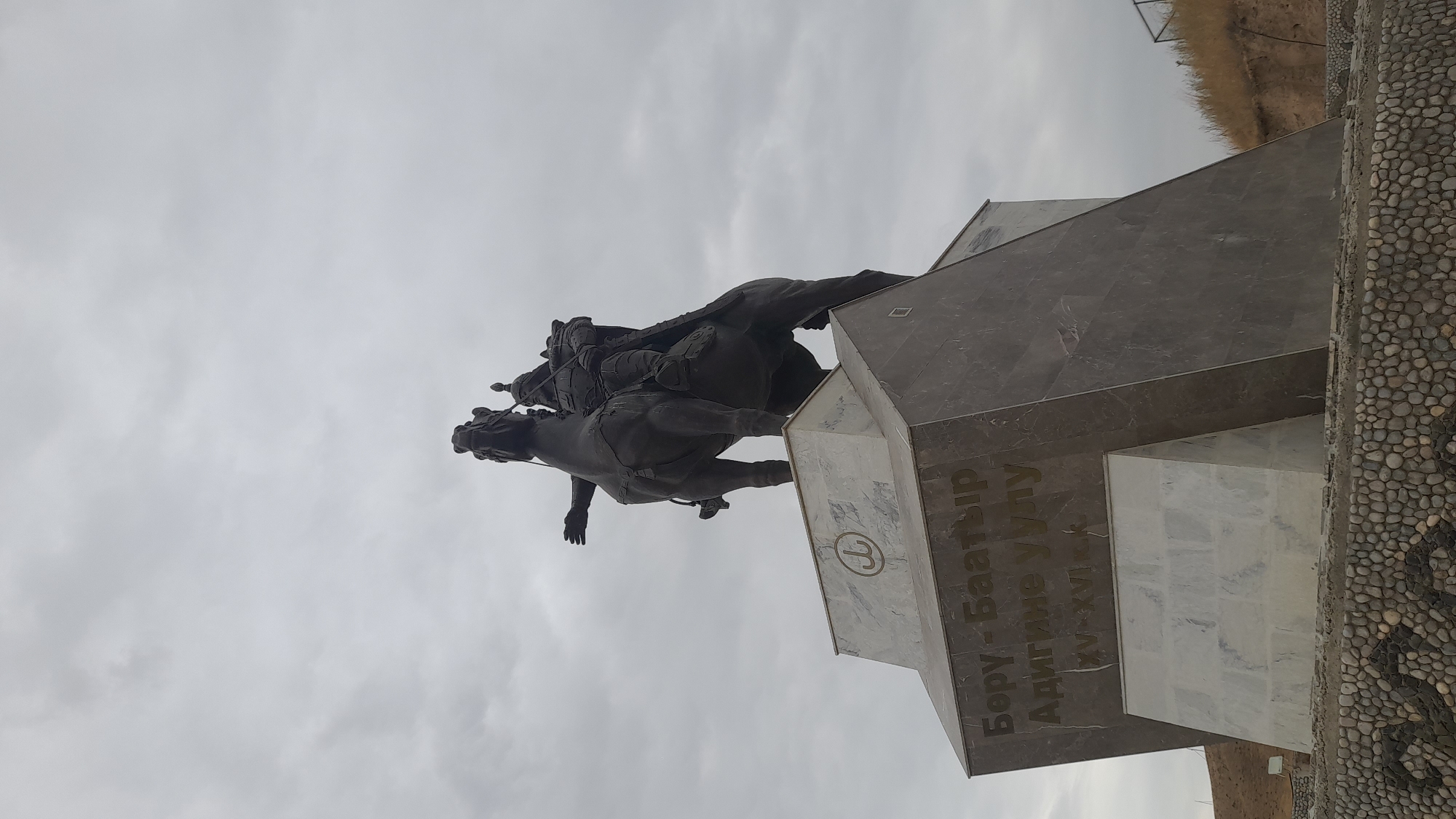 A low-angle outdoor shot shows a large bronze equestrian statue against an overcast sky. The statue depicts a person in armor on horseback, with their left arm outstretched. The monument is built on a multi-faceted stone pedestal with inscriptions. The base of the pedestal is decorated with a pattern of rounded stones. The background consists of dry vegetation on a hillside and a metal fence.
