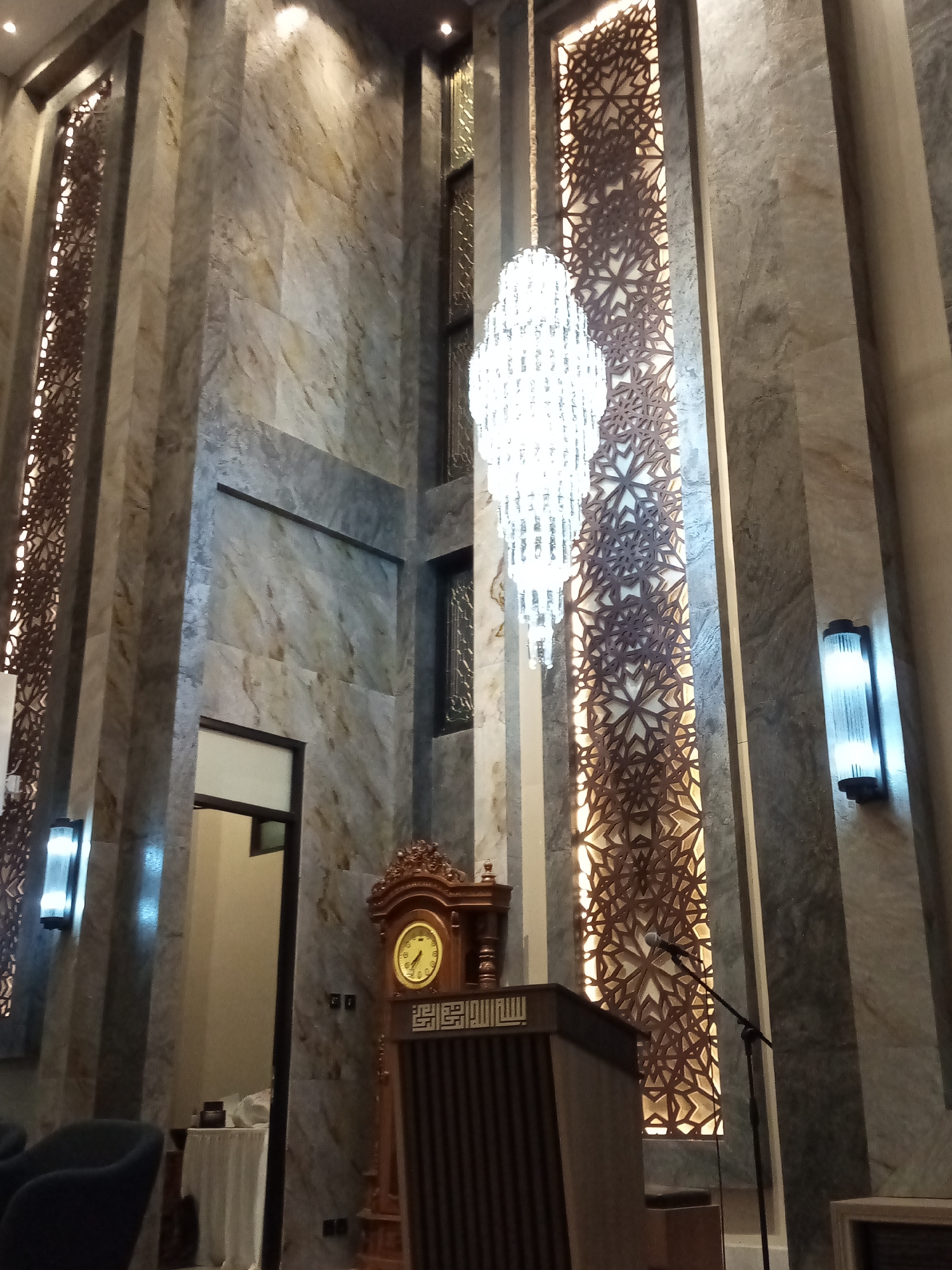The image depicts the interior of a building, likely a mosque or a similar place of worship, with a tall, marble-clad wall as the prominent feature. The architecture is modern with strong vertical lines created by the marble columns and decorative paneling.
The central element is a large, ornate chandelier hanging from the ceiling. To the right of the chandelier, a tall, decorative panel with an intricate geometric pattern is illuminated from behind, creating a warm glow. This panel extends vertically along the wall.
To the left of the chandelier, another similar decorative panel is visible. On the wall to the right, a modern wall-mounted light fixture with a rectangular blue glow is present.
In the foreground, a wooden podium with a microphone stand in front of it is positioned. To the left of the podium, a grandfather clock with a round face and ornate carvings is visible. Adjacent to the clock, a doorway leads to a room with a table covered by a white cloth.
The floor is not clearly visible in detail, but the overall impression is one of grandeur and careful design, common in places of religious significance. The lighting is a mix of ambient and directed light, highlighting the architectural features.
The image was taken in the city of Cihanjuang, Indonesia.