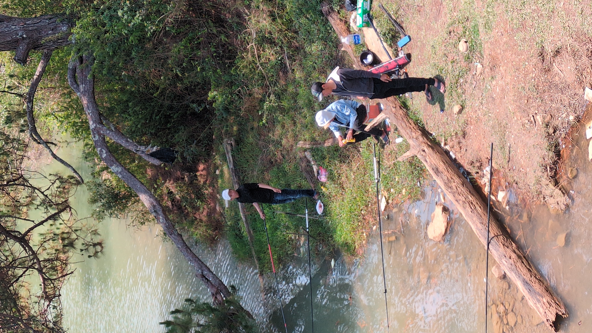 Three people are fishing by a river. The river is calm and shallow. A fallen tree lies partially in the water. Fishing rods are visible near the water's edge. The surrounding area is grassy with trees and vegetation. Based on the appearance of the landscape, this is likely in Lang Biang - Da Lat, Vietnam.
