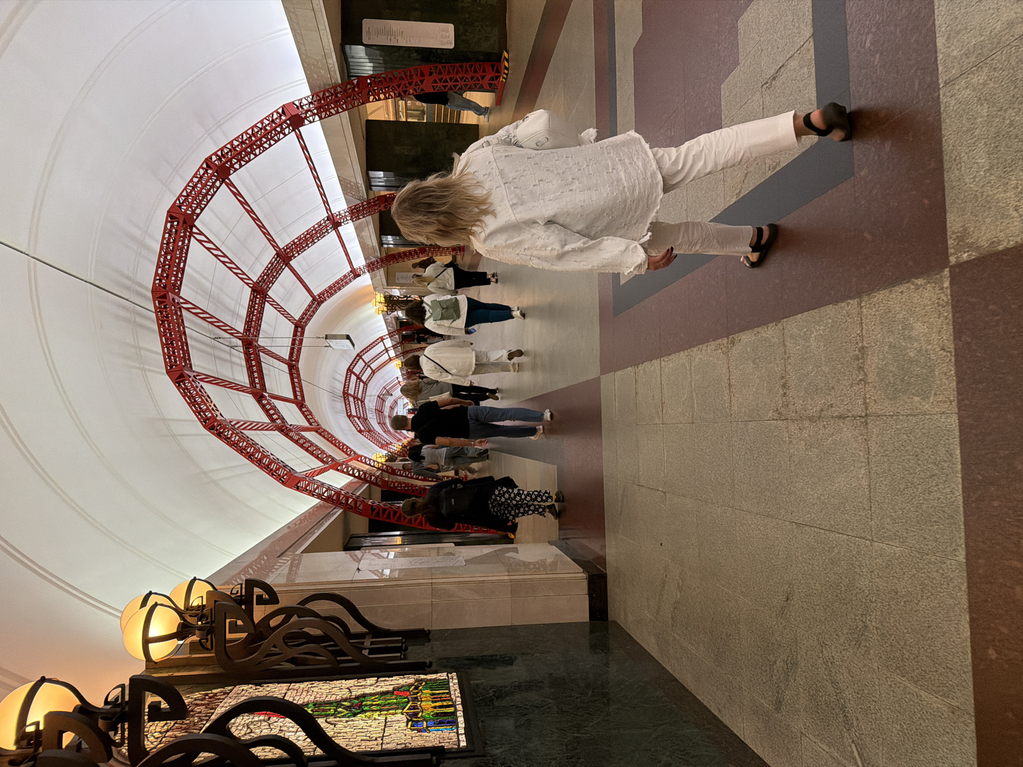 The image depicts an indoor architectural space, possibly a subway station or a grand hallway, with a distinctive red, latticed archway spanning the ceiling. The ceiling is white and curved, illuminated by warm yellow lights mounted on ornate, dark metal fixtures on the left side of the image. These fixtures also hold what appear to be stained-glass panels with colorful geometric patterns.
The floor is tiled with large, light-colored stone tiles, bordered by a band of dark red and a darker grey stripe. Several people are walking away from the viewer down a corridor. The person closest to the camera, in the foreground, is a woman with blonde hair wearing a loose-fitting white shirt and white pants, with black sandals. She is carrying a white handbag. Other individuals in the mid-ground are also walking away, dressed in casual attire. One woman has a green backpack, and another is wearing patterned pants. In the background, a train can be seen through an opening on the right. The overall impression is of a well-lit, spacious, and decorated public area.