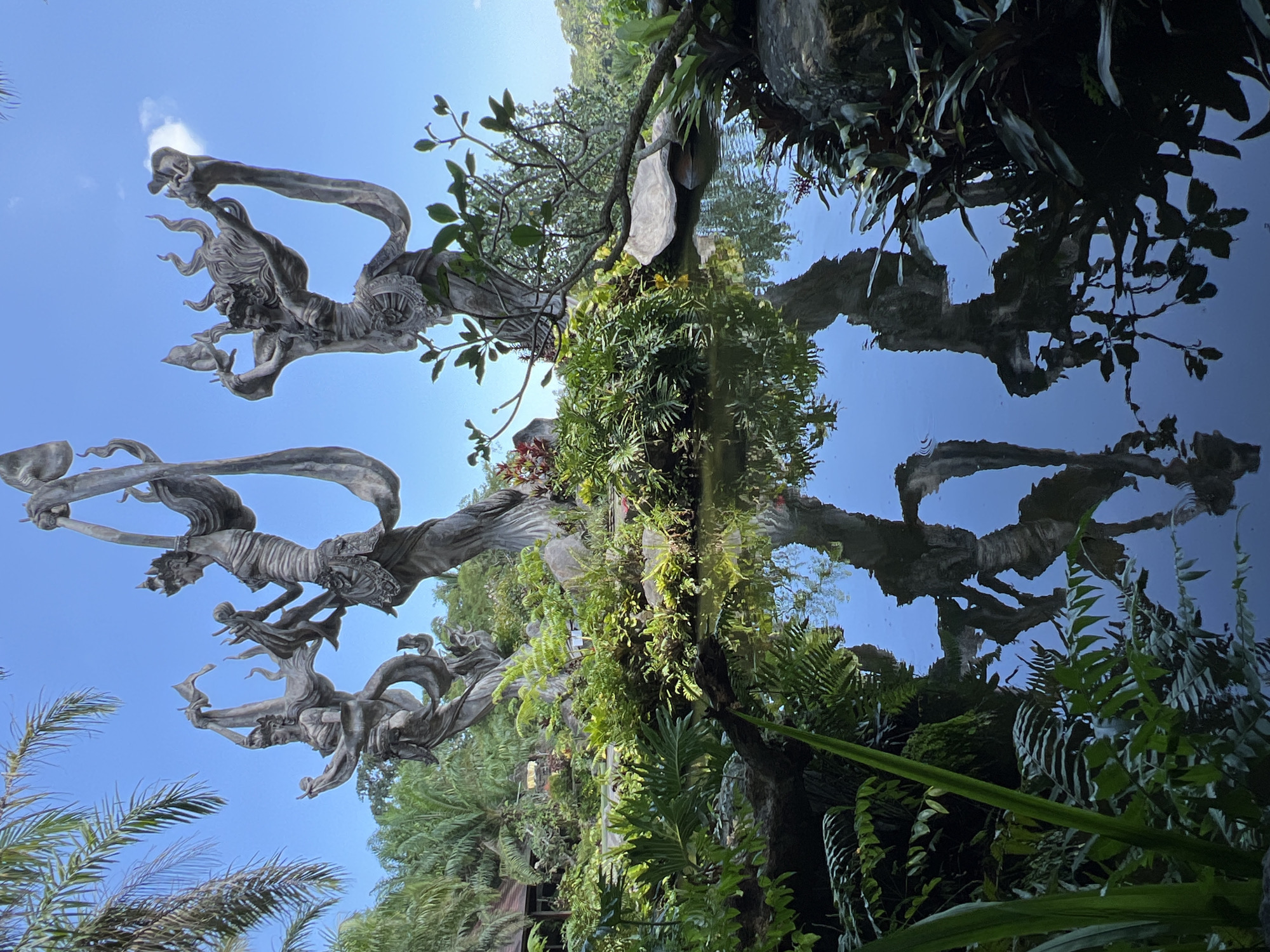 The image shows an upside-down view of what appears to be a sculpture depicting figures, possibly deities, against a blue sky. The sculpture is grayish in color and intricate in detail. The lower portion of the image is dominated by lush green vegetation, including trees, ferns, and various plants. The upper portion shows what seems to be the reflection of the sculptures and vegetation in a pool of water, resulting in a mirror-image effect. The overall scene is likely located in a tropical or subtropical environment. It is in Kedewatan, Indonesia.