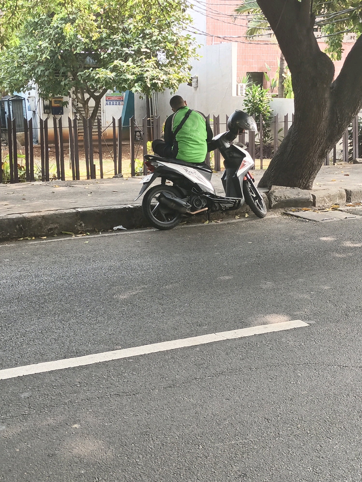 A person wearing a green vest sits on a scooter, parked on the side of a road in South Jakarta, Indonesia. The scooter is white and black, with a black helmet placed on the handlebars. The person is facing away from the camera, and their legs are extended. The scooter is parked on a concrete curb, with a sidewalk behind it. A fence made of dark wood is visible behind the sidewalk, with trees and buildings in the background. The road is paved with asphalt and has a white line painted on it. The scene appears to be a typical urban street with moderate traffic.