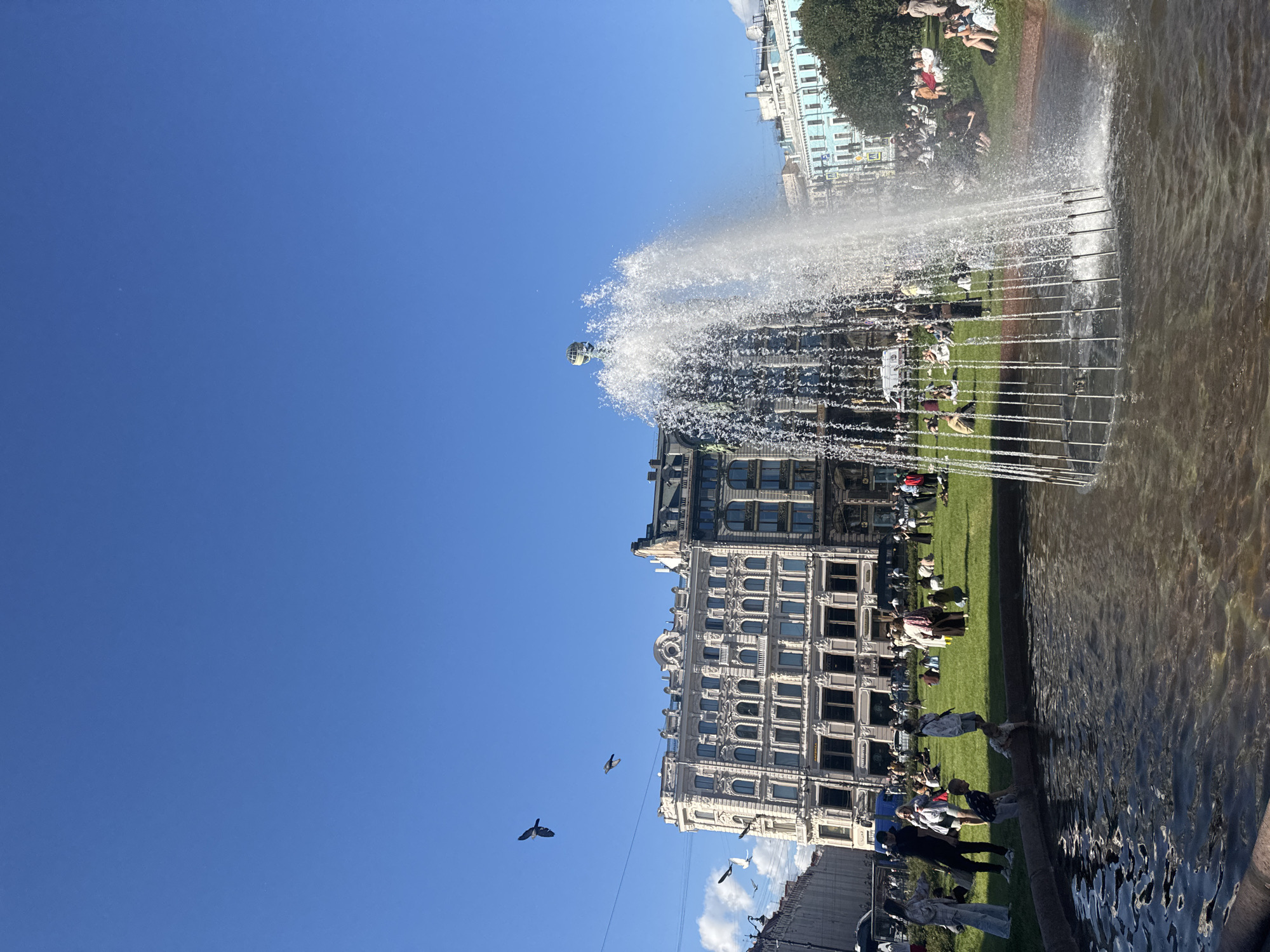 This is a photograph of a fountain with a large building and people in the background, set against a clear blue sky. The image is oriented vertically, with the sky occupying the top left portion and the fountain and building filling the right and bottom sections.
The central subject is a wide, multi-tiered fountain spraying water high into the air. The water forms a misty curtain, and some of the droplets catch the sunlight, creating a subtle rainbow effect. In the background, several people are visible, some lounging on a grassy area, others walking or standing. The people are dressed in casual summer clothing.
Behind the fountain and the people is a large, ornate building with a classical architectural style. It has a light-colored facade, numerous windows, and decorative details. To the far left of the frame, another building with a lighter blue facade and more modern architectural elements can be seen. A tree with green foliage is also present. Several birds are flying in the sky.
The scene appears to be set during the daytime, likely a sunny afternoon, given the bright lighting and clear sky. The presence of people enjoying the outdoors suggests pleasant weather. The location cues, such as the architectural style of the buildings and the general atmosphere, are consistent with an urban setting in Saint Petersburg, Russia. The overall mood is relaxed and summery, with people enjoying a public space on a fine day. There is no visible text in the image.
