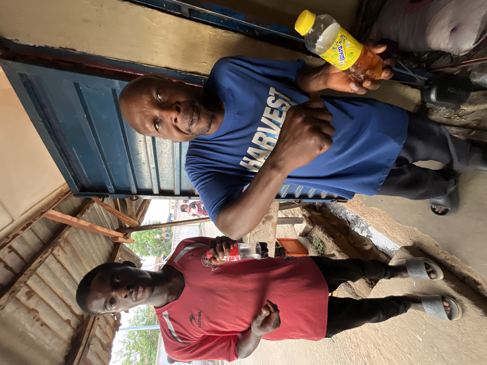 Two men are standing in what appears to be a partially covered outdoor space. The man on the left wears a red jersey with the word "ASTORE" visible, while the man on the right wears a blue t-shirt with the word "HARVEST" printed on it. The man in the red jersey holds a Coca-Cola bottle, and the man in the blue shirt holds a bottle of a yellow liquid. Both men are looking at the camera. They are standing on a cement or concrete surface. Overhead, there is a roof structure of corrugated metal and wooden supports. The location is Jos, Nigeria.