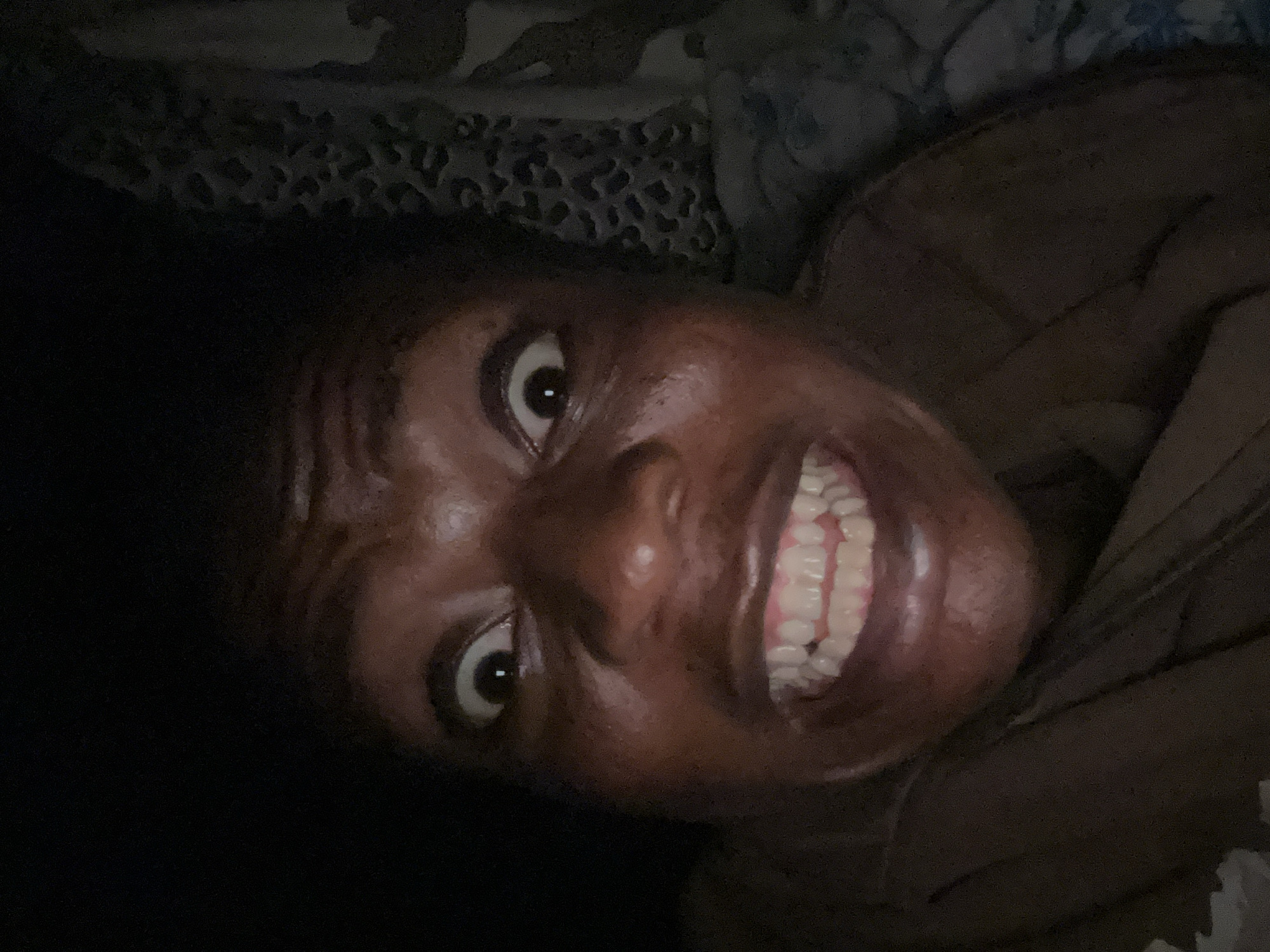 A close-up, tilted portrait shows a person with dark skin, wide eyes, and a broad grin revealing their teeth. The person is lying down. A patterned fabric is visible near the top of the image, suggesting a blanket or bedding. The environment appears dark.