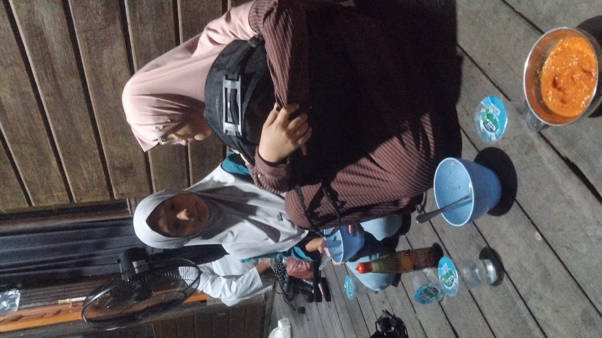 Two young women wearing hijabs are sitting on a wooden deck. The woman on the left wears a light grey hijab and a blue shirt, and she is looking down at a light blue bowl in front of her. The woman on the right wears a pink hijab and a brown and white striped shirt. She is looking to her left, away from the camera, and has a black backpack in front of her. There are several small plastic cups and bowls on the deck, as well as a bottle of sauce. A fan is visible in the background to the left. The setting appears to be outdoors, possibly at night, given the lighting. The location is Bangun Rejo, Indonesia.
