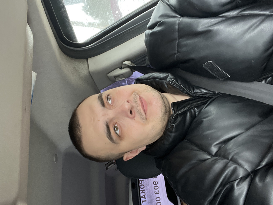A male individual is observed seated inside the passenger compartment of a vehicle in Rogachovo, Russia. He appears to be an adult, exhibiting short dark hair, light brown eyes, and light facial stubble. His gaze is directed upwards and to the right, away from the camera, with a neutral expression and a slight upturn at the corners of his mouth. He is attired in a black, shiny, puffer-style jacket, secured by a visible black seatbelt diagonally across his torso and left shoulder. The vehicle's interior features a light gray headliner and trim. Through the passenger window on the right, a blurry outdoor scene consistent with overcast or winter weather is visible, showing indistinct trees or foliage. On the left side of the frame, partially obscured and reflected, is Cyrillic text, appearing inverted as "ТАКОР" (likely part of "ПРОКАТ" - Rental) and numbers "000 ε0ε" (likely "303 000"), suggesting an external commercial establishment. The scene depicts the individual in a stationary or moving vehicle.