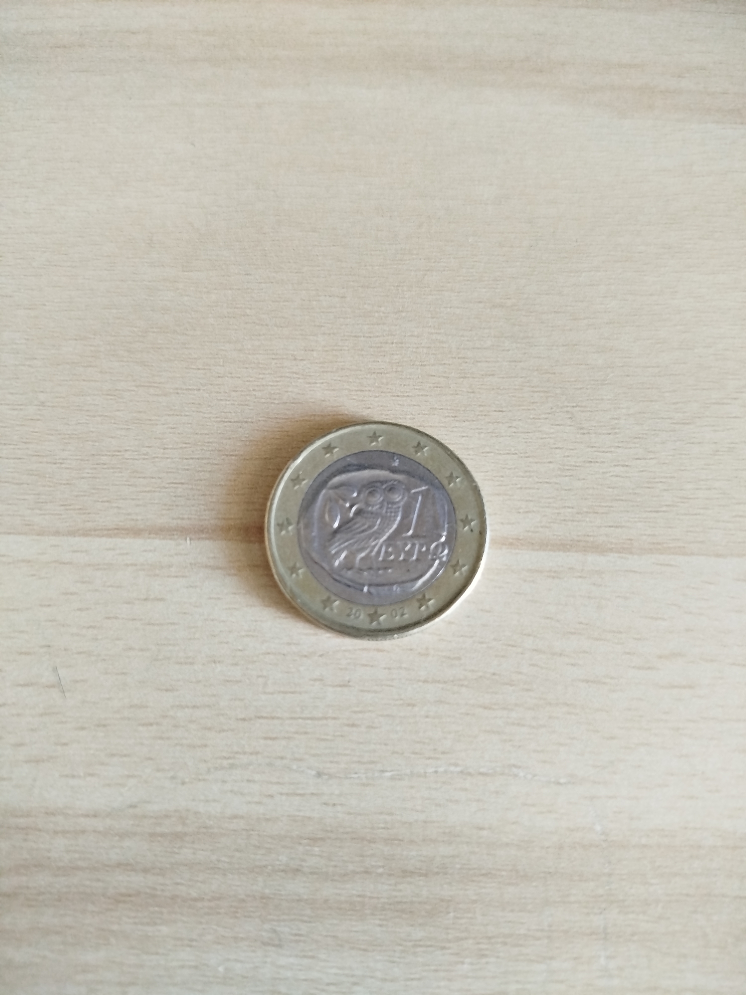 A single 1 Euro coin rests on a light brown wooden surface. The coin displays the image of an owl and the word "EURO". The coin is oriented with the owl's head pointing towards the top of the image and the word "EURO" oriented horizontally. The coin's outer ring is gold with twelve stars and the year "2002" inscribed at the bottom. There are no people, subjects, or other objects in the immediate vicinity. The environment appears to be indoors based on the even lighting and the surface the coin is resting on. The location is not verifiable as Varna, Bulgaria based on the image provided.