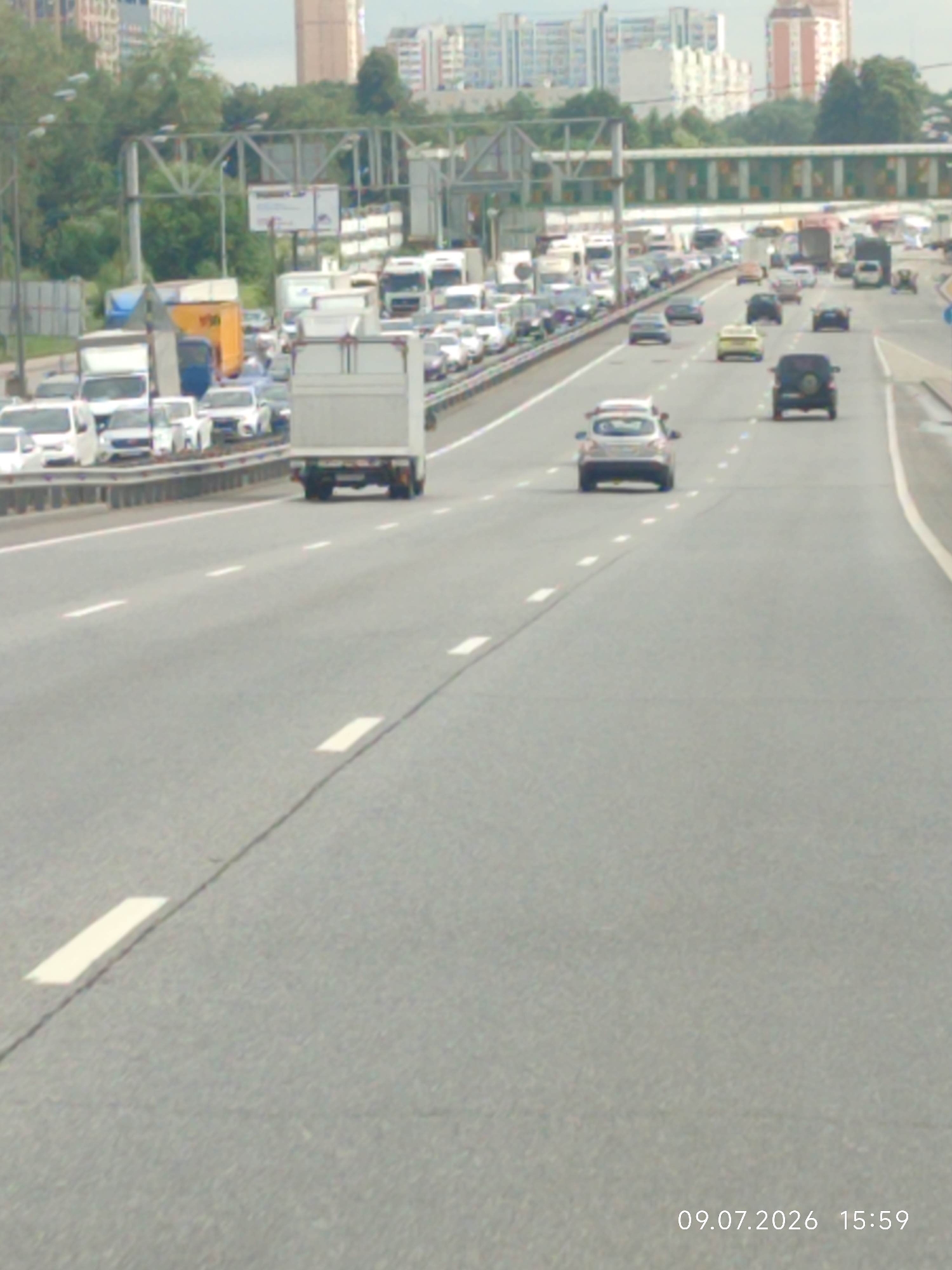 The image shows a traffic jam on a multi-lane highway with buildings and trees in the background. The road is asphalt, with white lane markings. There are numerous vehicles on the highway, including cars, trucks, and vans, all moving slowly or stopped. The sky is overcast. A timestamp in the lower right corner indicates the date and time as 09.07.2026 15:59. The location is identified as Vinogradovo, Russia.
