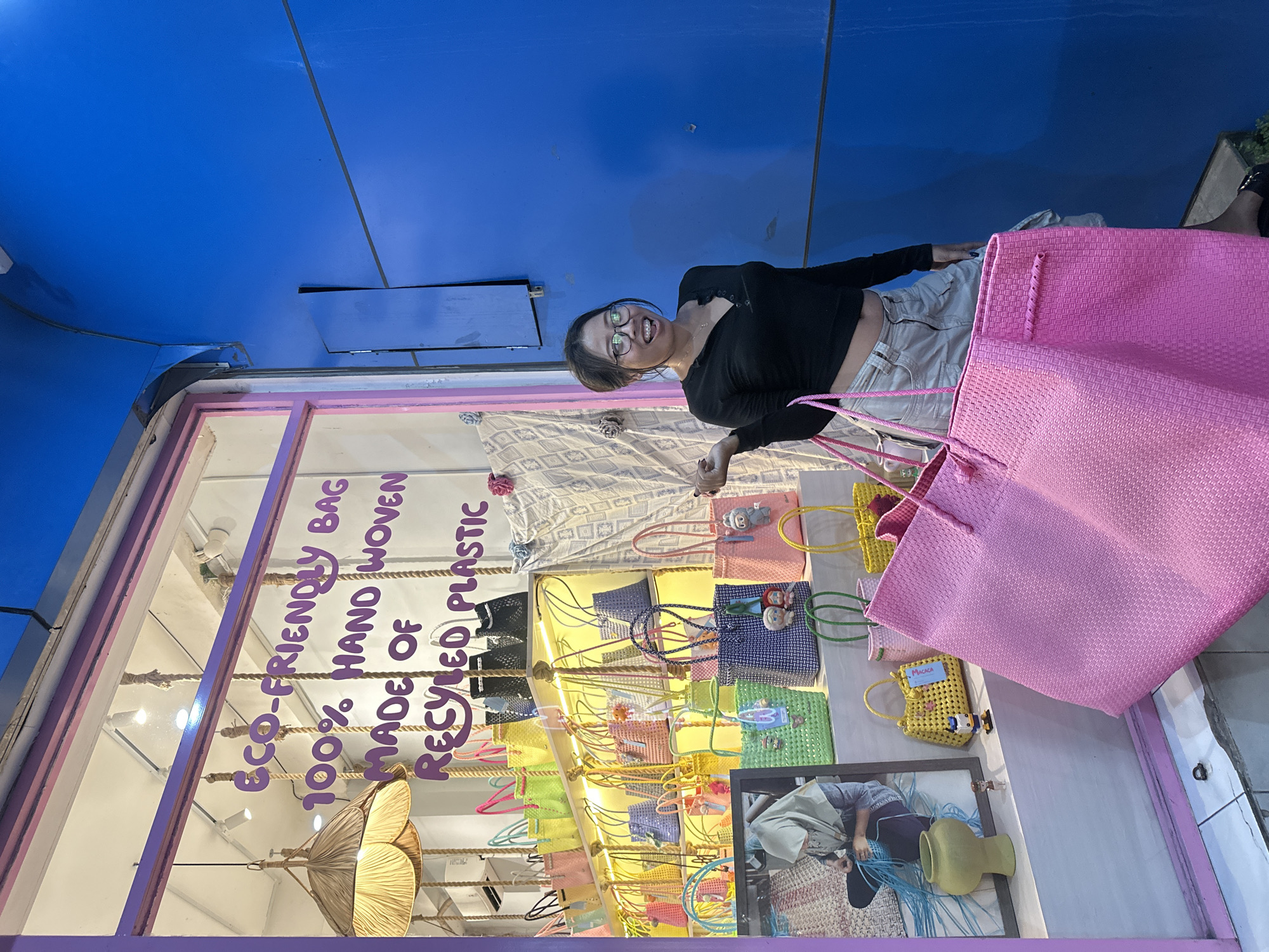 A woman stands in front of a storefront in what appears to be Ubud District, Indonesia. The window display has signs indicating that the items are "Eco-Friendly Bag 100% Hand Woven Made of Recycled Plastic." The store displays various colorful hand-woven bags. The woman is holding a large pink bag. The building has a blue ceiling. The woman wears glasses and a black shirt.