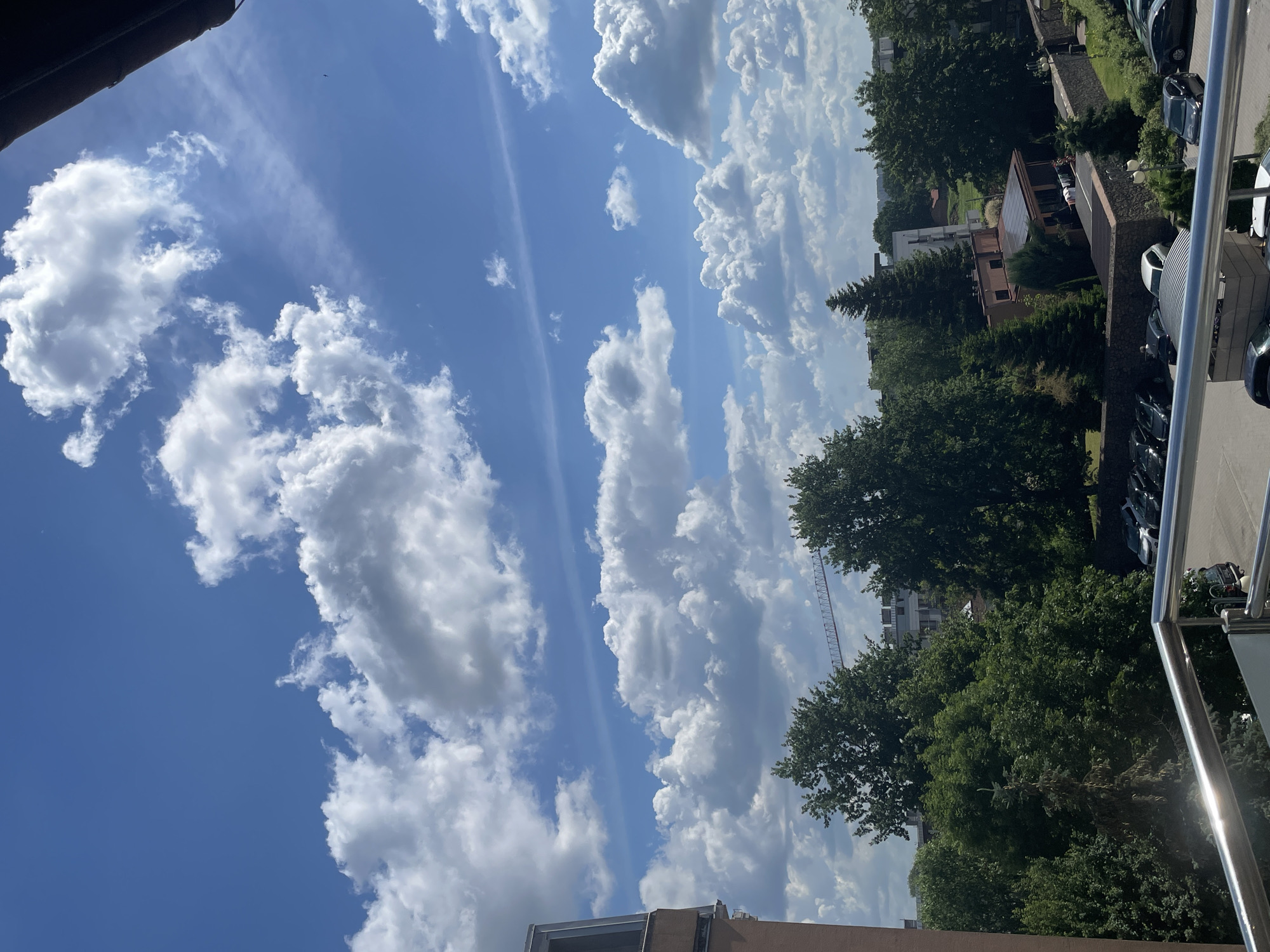 The image shows a cloudy sky over a city. The sky is blue with numerous white and gray clouds, some of which are fluffy cumulus clouds. A sliver of a dark structure, possibly an awning or roof edge, is visible in the upper right corner. Below the sky is a cityscape with buildings and trees. In the foreground, a metal railing is visible, suggesting the photo was taken from a balcony or elevated vantage point. A building with a brown facade is on the left. A construction crane is visible in the background. Several trees with green foliage are spread throughout the scene. A parking lot with several cars is visible in the lower part of the image. A stone wall borders a grassy area with a building that has solar panels on its roof. The overall scene depicts an urban environment on a partly cloudy day.