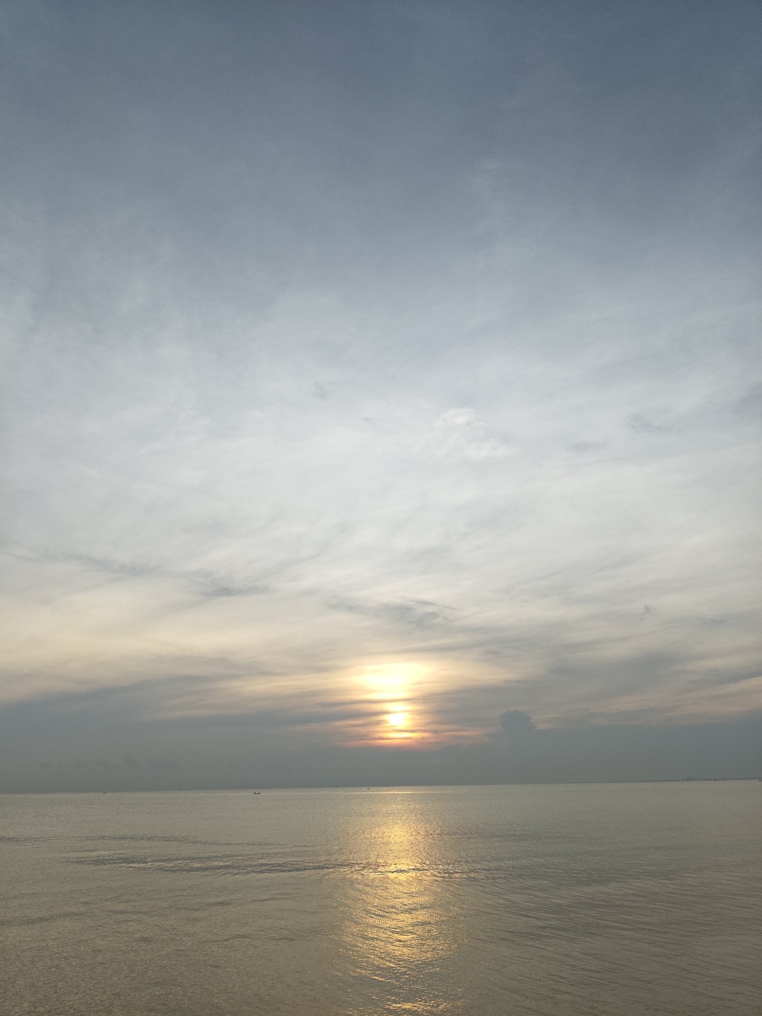 The image depicts a sunrise or sunset over the sea, likely in Lhokseumawe, Indonesia. The sun is positioned low on the horizon, with its light reflecting on the calm water surface. The sky is predominantly grey with white clouds. The horizon is mostly free of structures, there are tiny, dark shapes that may be boats.