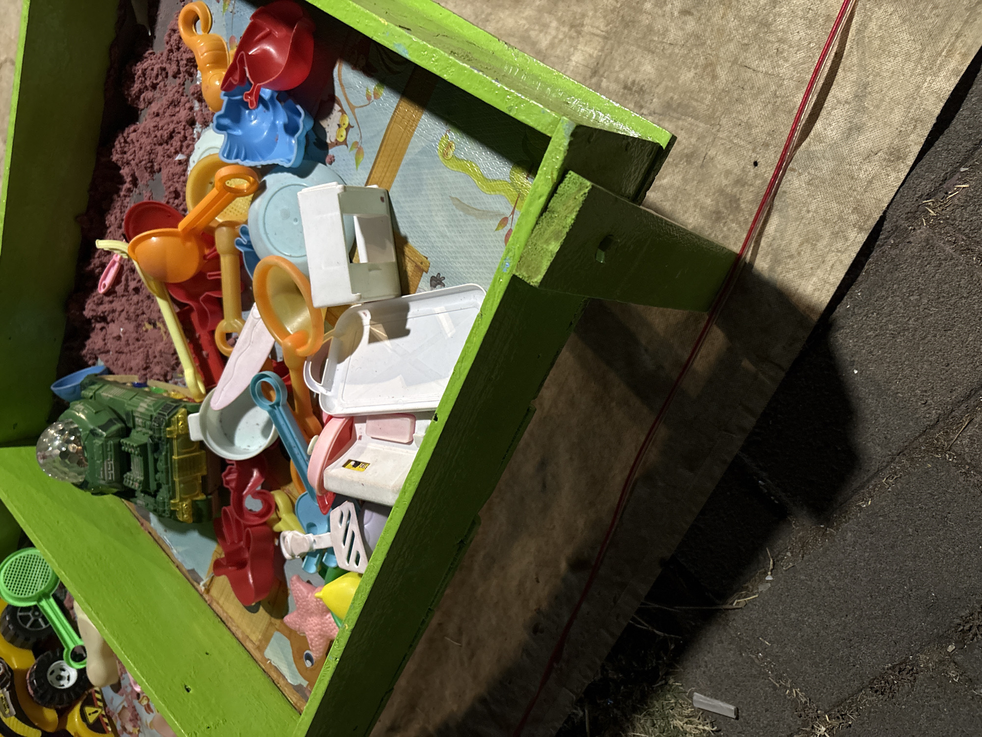 The image shows a close-up, high-angle view of a green sandbox filled with various toys and kinetic sand. The sandbox appears to be made of wood, with a rough texture on its outer surface, particularly on the legs. Inside the sandbox, a variety of plastic toys are scattered amongst the reddish-brown sand. These include a green toy tank with a disco ball on top, multiple scoops and shovels in different colors (orange, blue, pink, yellow), a pink starfish-shaped mold, a yellow lemon-shaped mold, and a white toy house. The base of the sandbox is lined with a mat featuring cartoonish animal illustrations, including an owl and a snake. In the background, part of a yellow toy vehicle with black wheels is visible. The sandbox is placed outdoors, as indicated by the presence of a dark-colored surface resembling pavement or concrete in the lower left of the image, with some dried grass and debris scattered on it. A red wire runs horizontally across the bottom of the frame, partially obscured by shadows cast by the sandbox. The lighting suggests it may be evening or nighttime, with strong shadows. The location is identified as Palimanan, Indonesia.