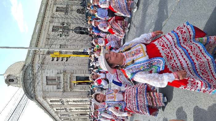 A procession of women in traditional Peruvian clothing walks along a street. They wear wide-brimmed white hats, embroidered white lace tops, red skirts with white and blue detailing. The background features a grand, old stone building with a domed tower, yellow traffic lights, and power lines. The group appears to be part of a cultural celebration.