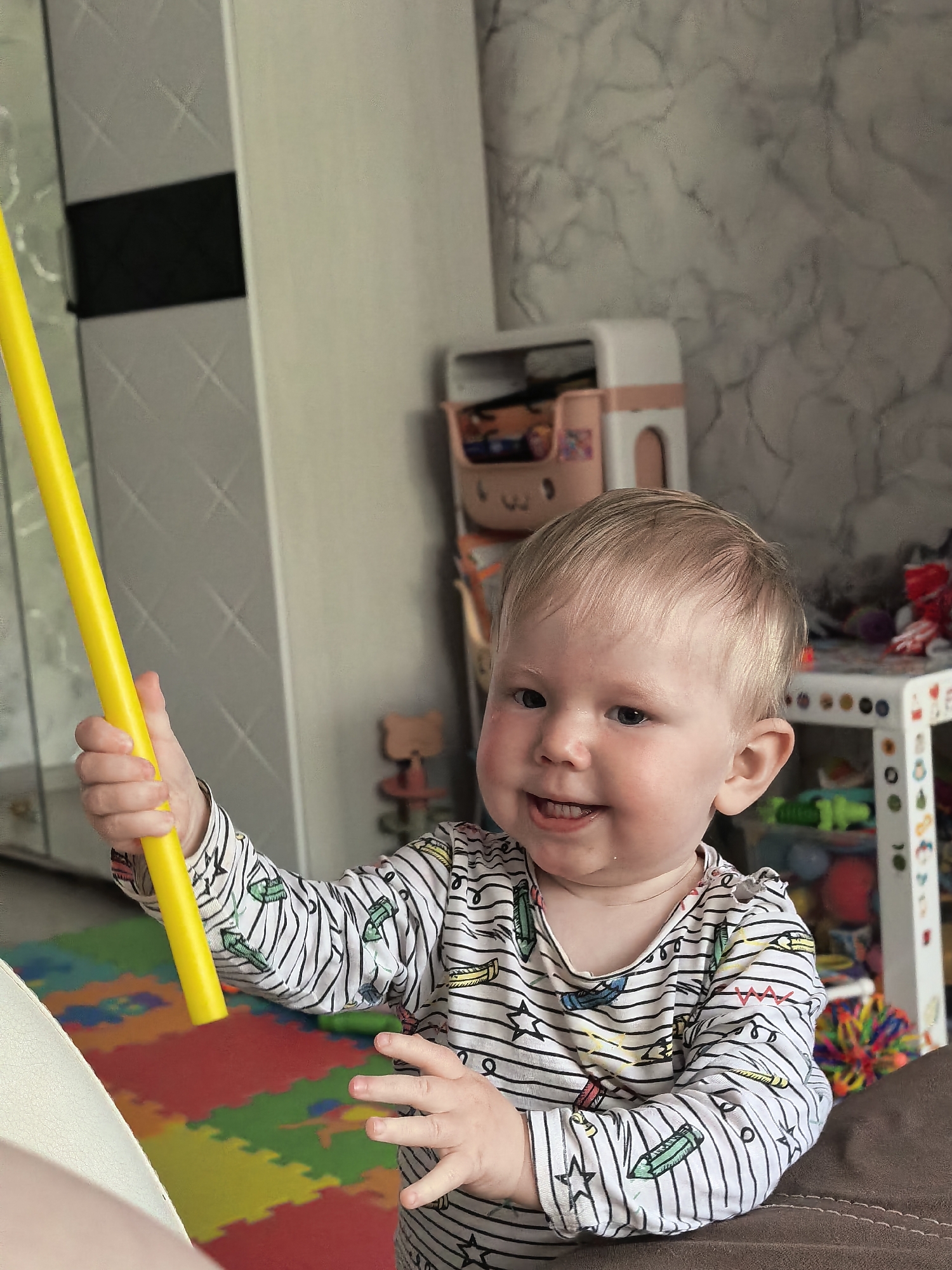 A baby is standing indoors. The baby is holding a long yellow stick. A patterned playmat is on the floor. Toys are on a shelf in the background. The baby is wearing a striped shirt with drawings of pencils and stars on it. The photo was taken in Sterlitamak, Russia.